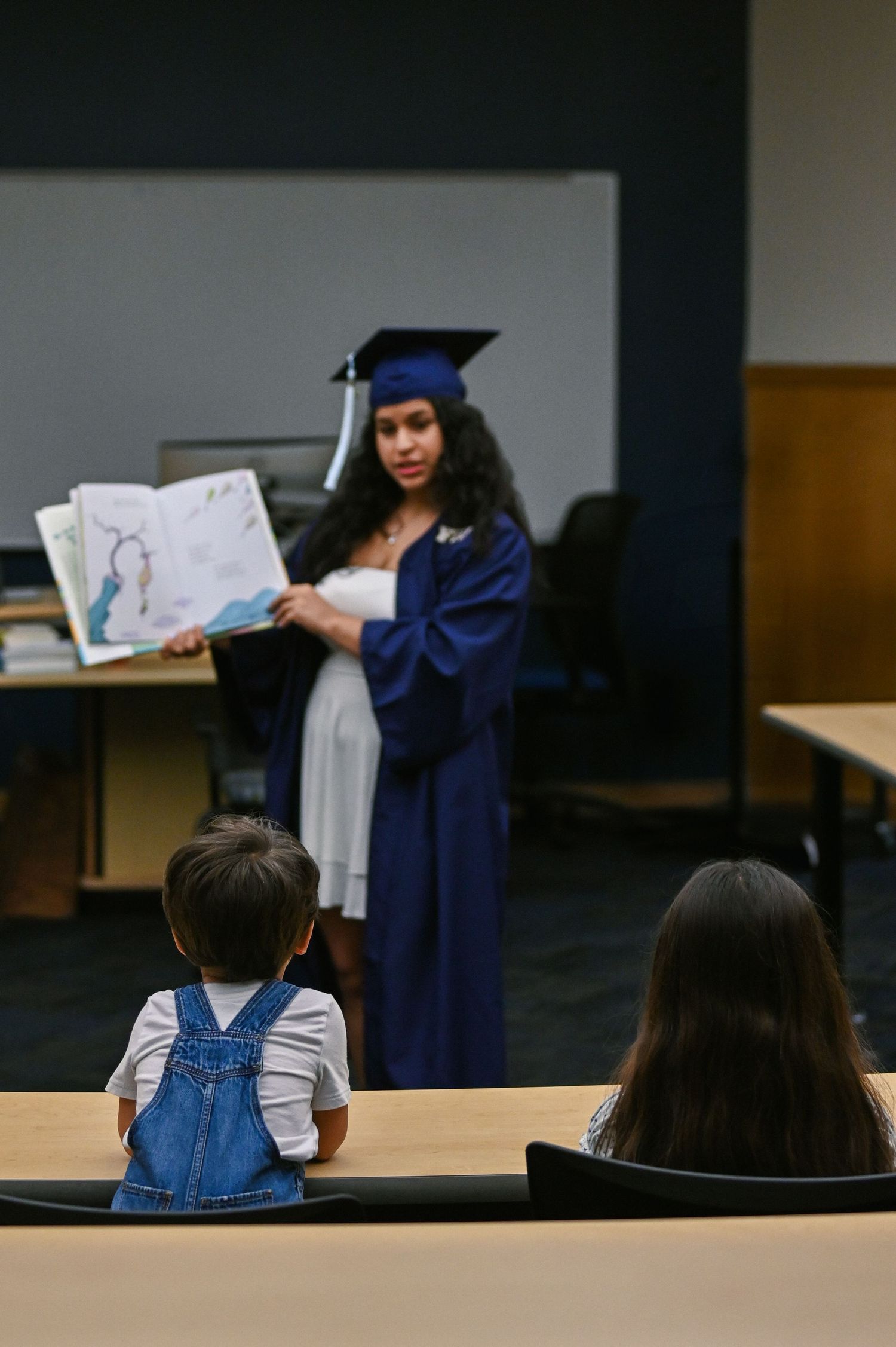 A graduate in blue cap and gown displays artwork in a classroom setting.