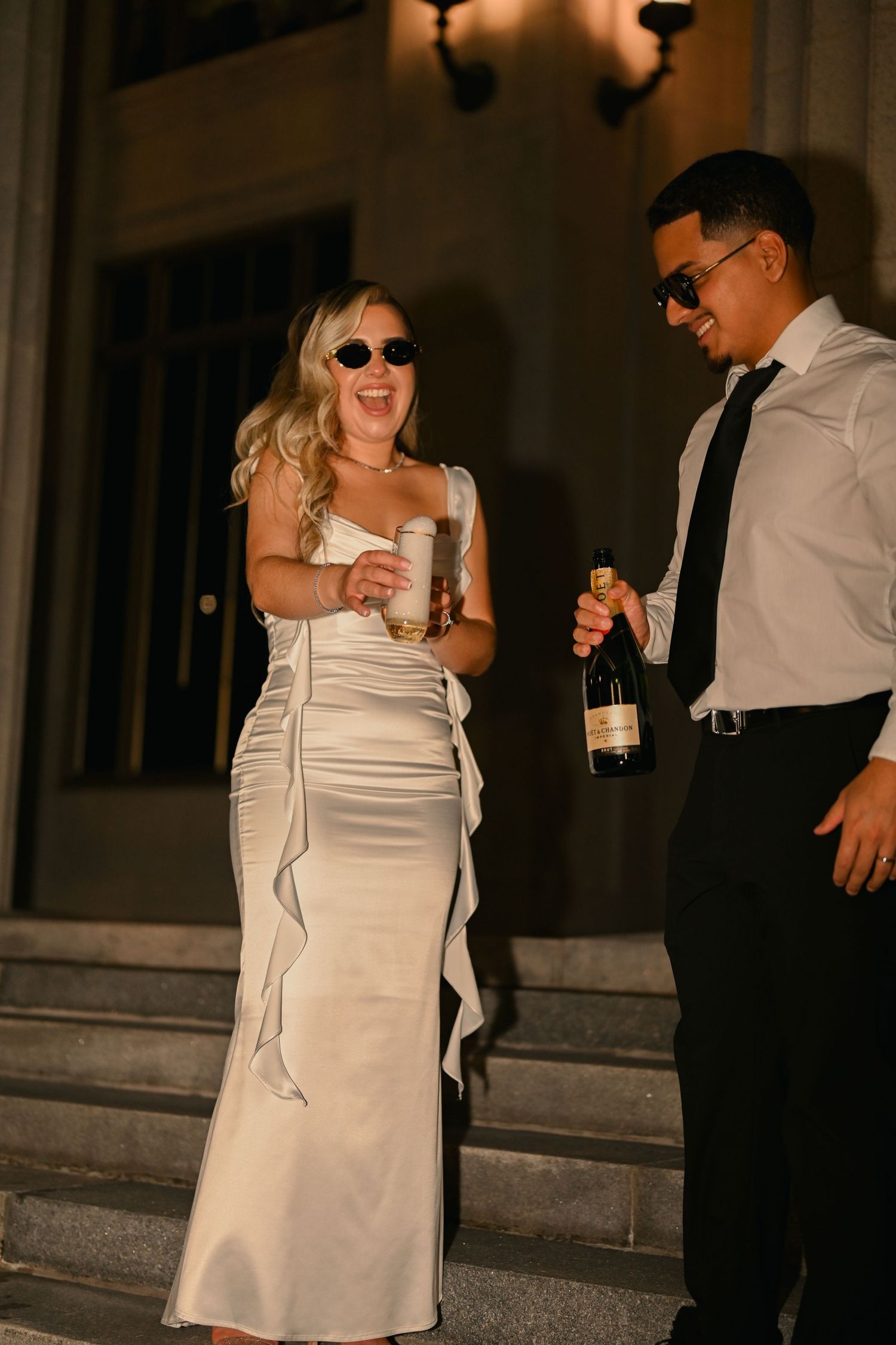 A couple in formal attire dances and celebrates at night, with the woman wearing a white dress and sunglasses.