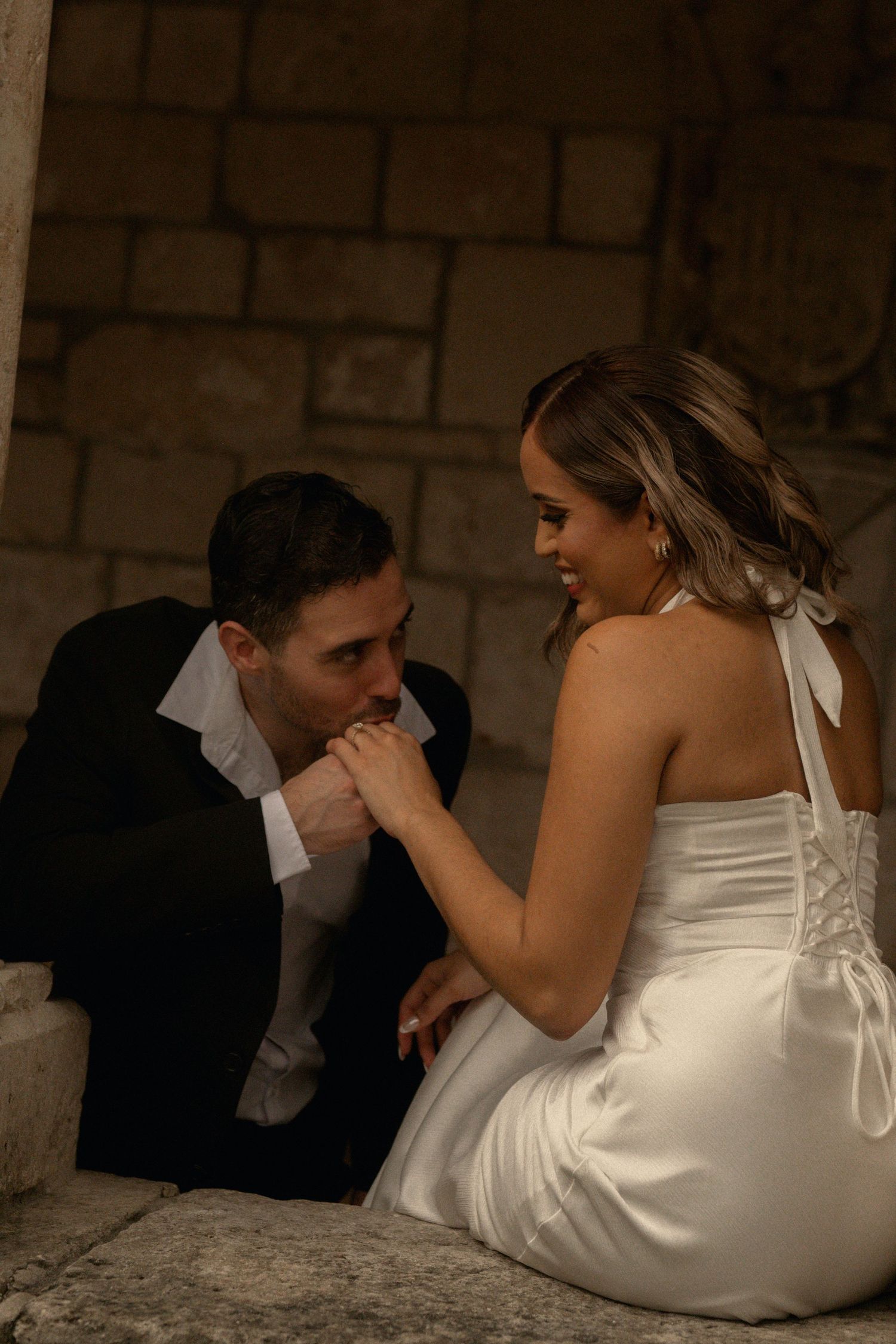 Groom in a dark suit kisses the hand of a smiling bride in a white satin dress while seated against a rustic stone wall.