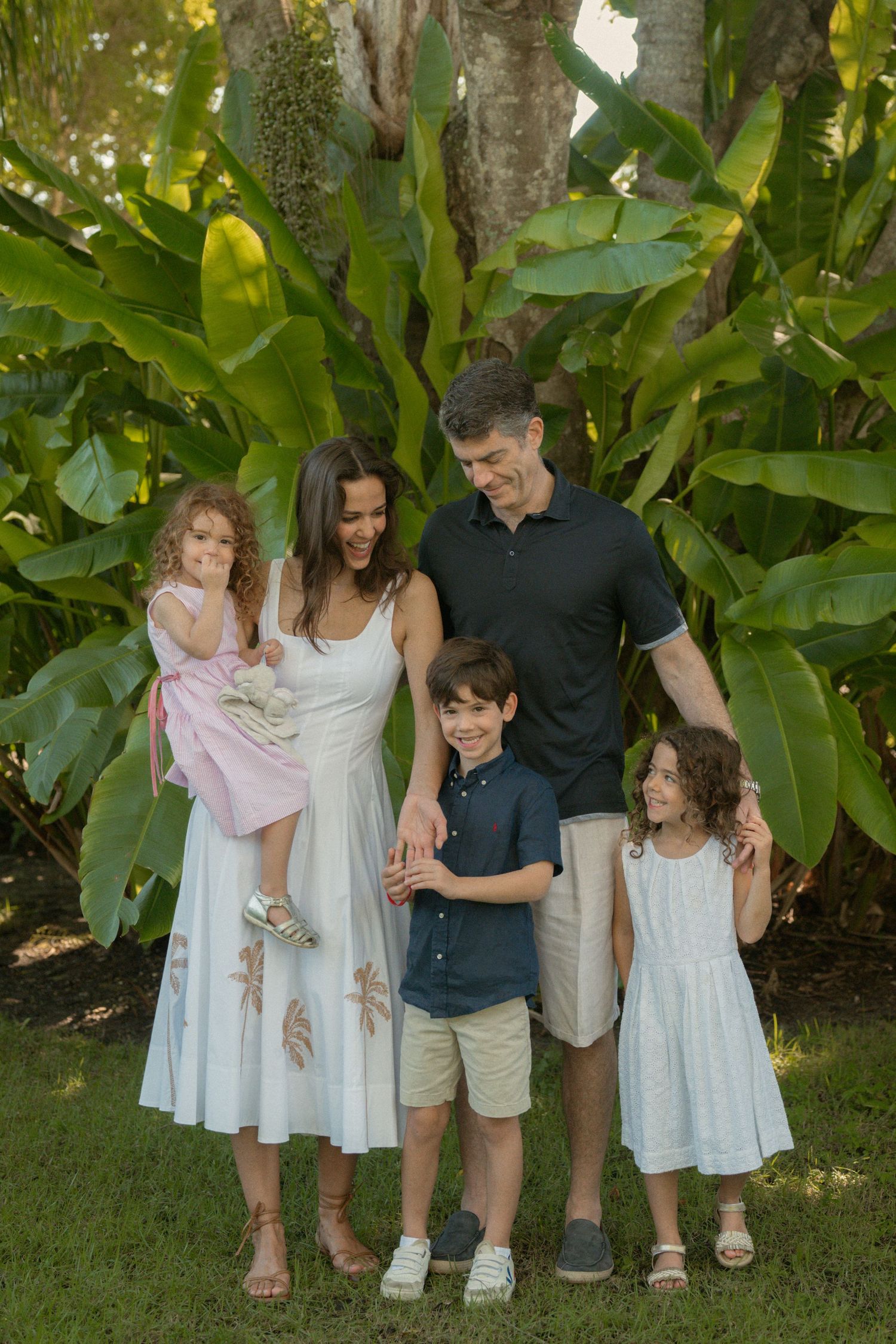 Smiling family of five in coordinated outfits poses together on a lush green lawn surrounded by tropical banana plants and foliage.