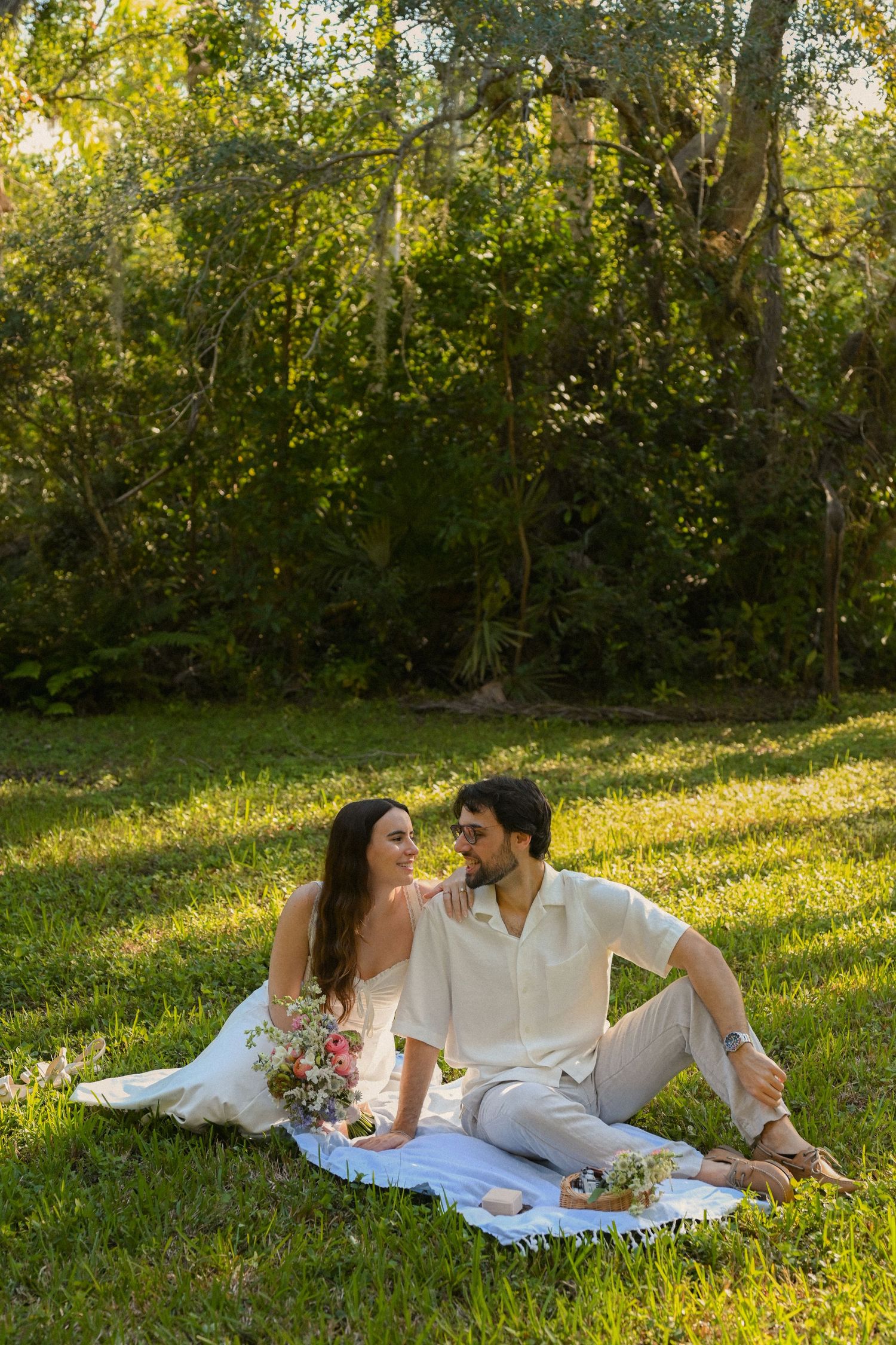 Couple having a picnic date while lounging on white blanket in sun-dappled park setting.