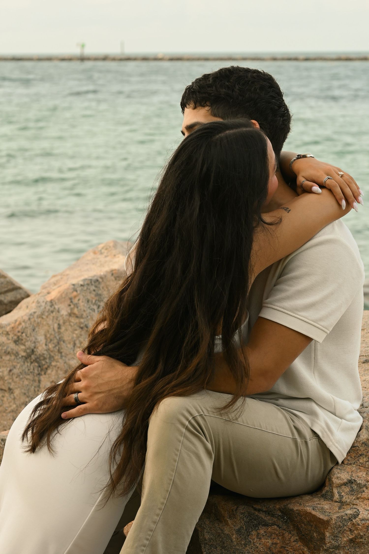 Couple in light colored clothing embracing on rocky shoreline during golden hour.
