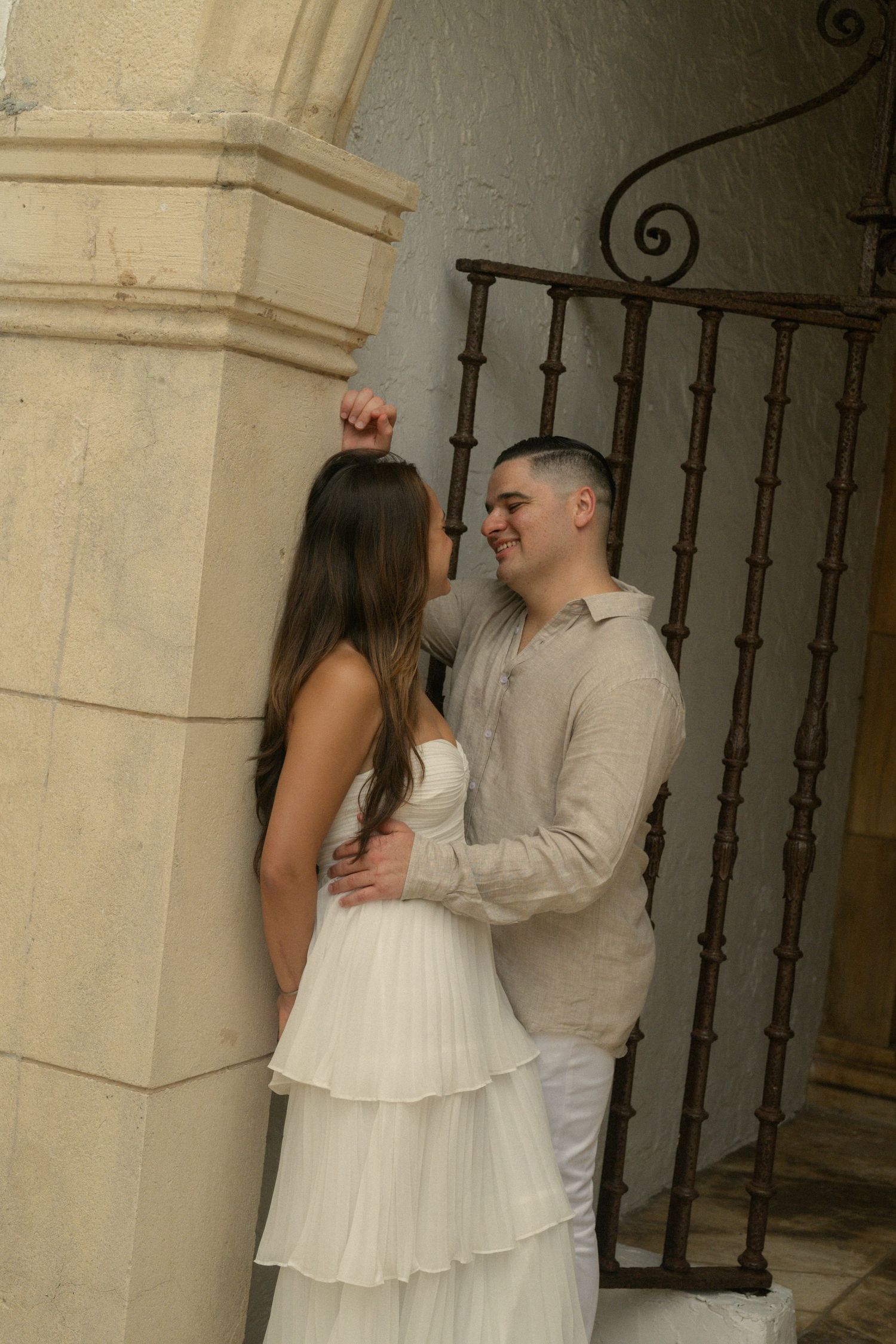 Romantic couple embracing near stone arch and ornate iron gate, woman in white tiered dress, man in linen shirt.