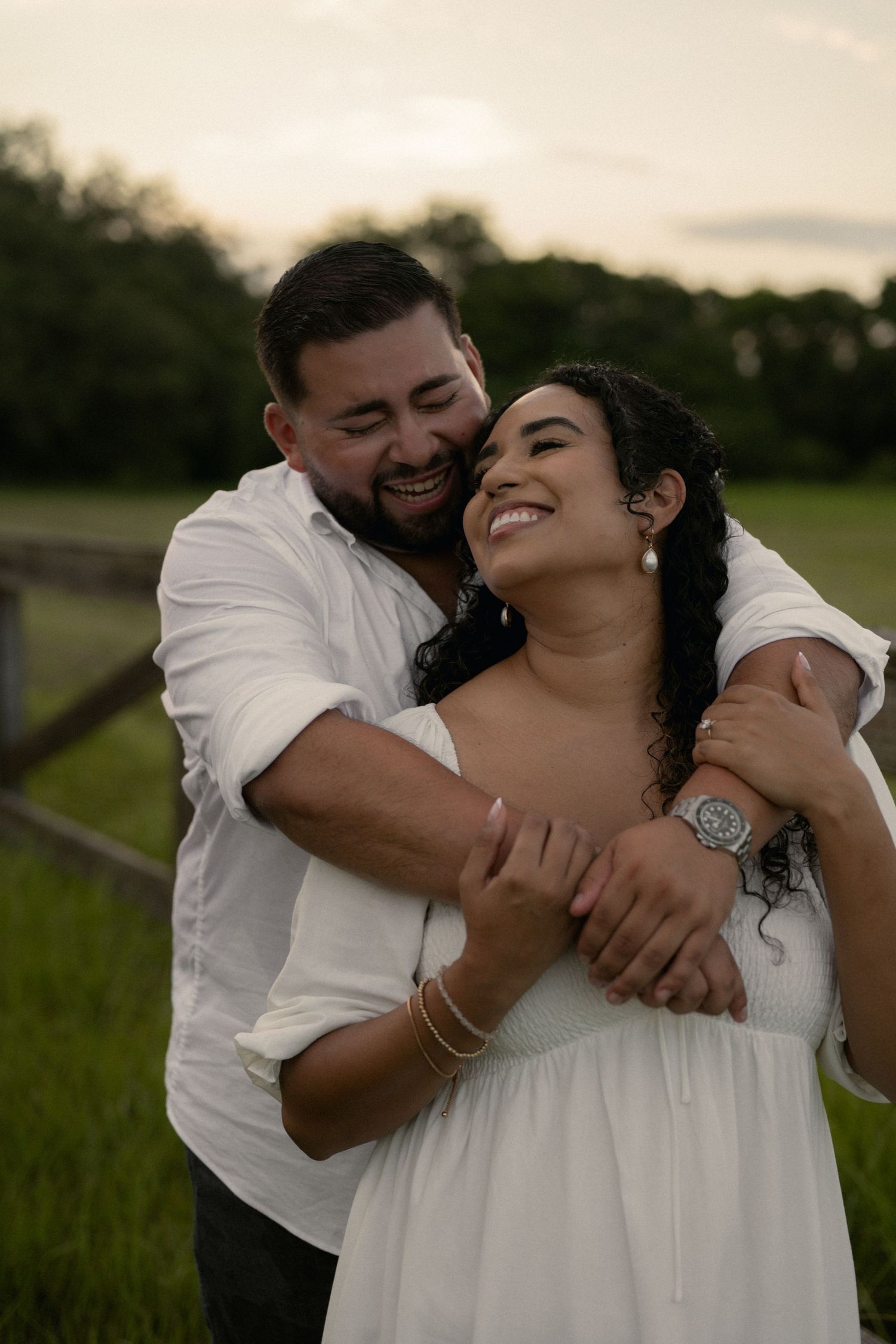 A smiling couple embraces outdoors at sunset, woman in white dress, man in white shirt, green field background.
