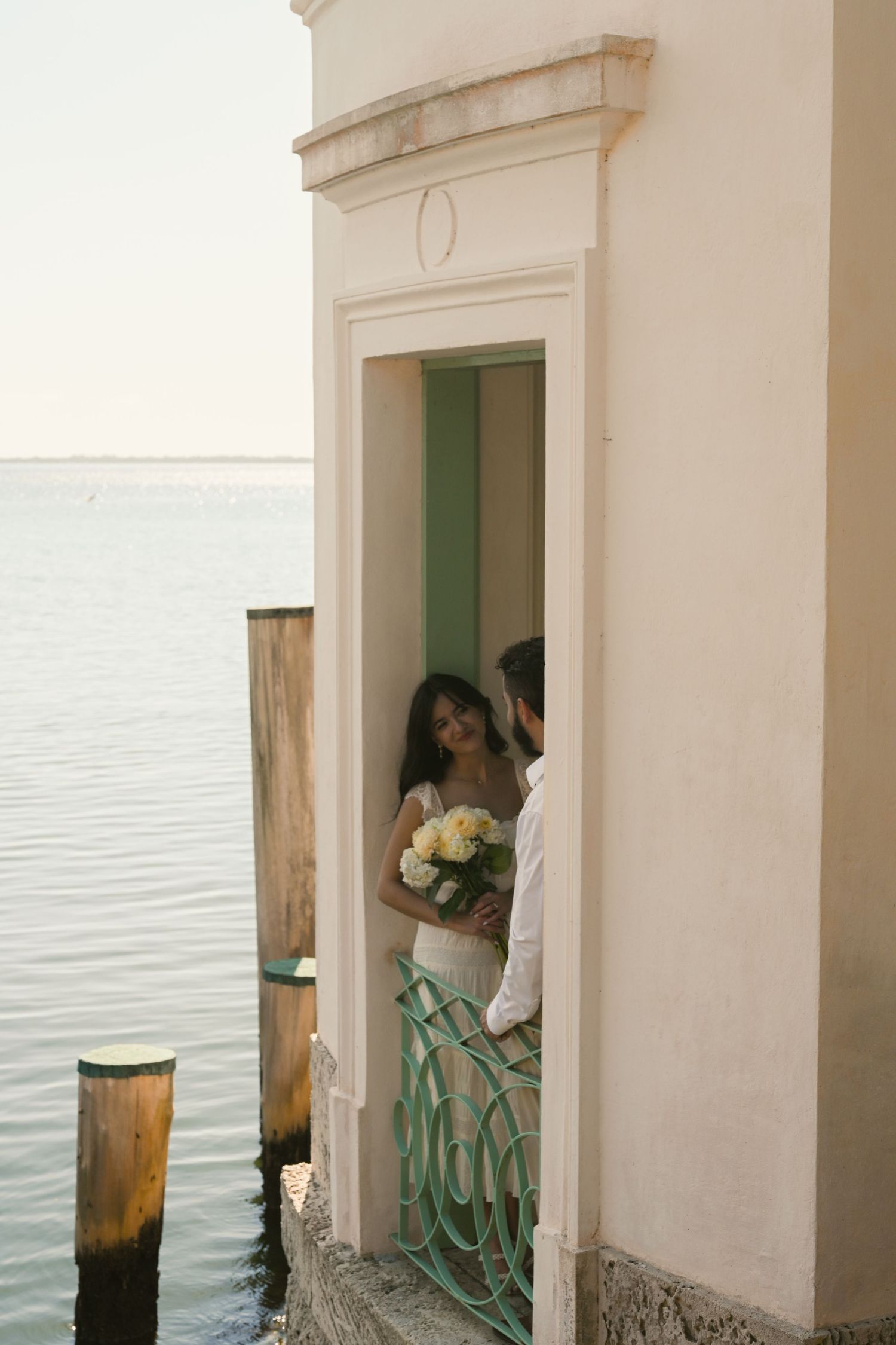 Bride and groom share a romantic moment in a white archway doorway overlooking serene lake waters, bride holding white bouquet.