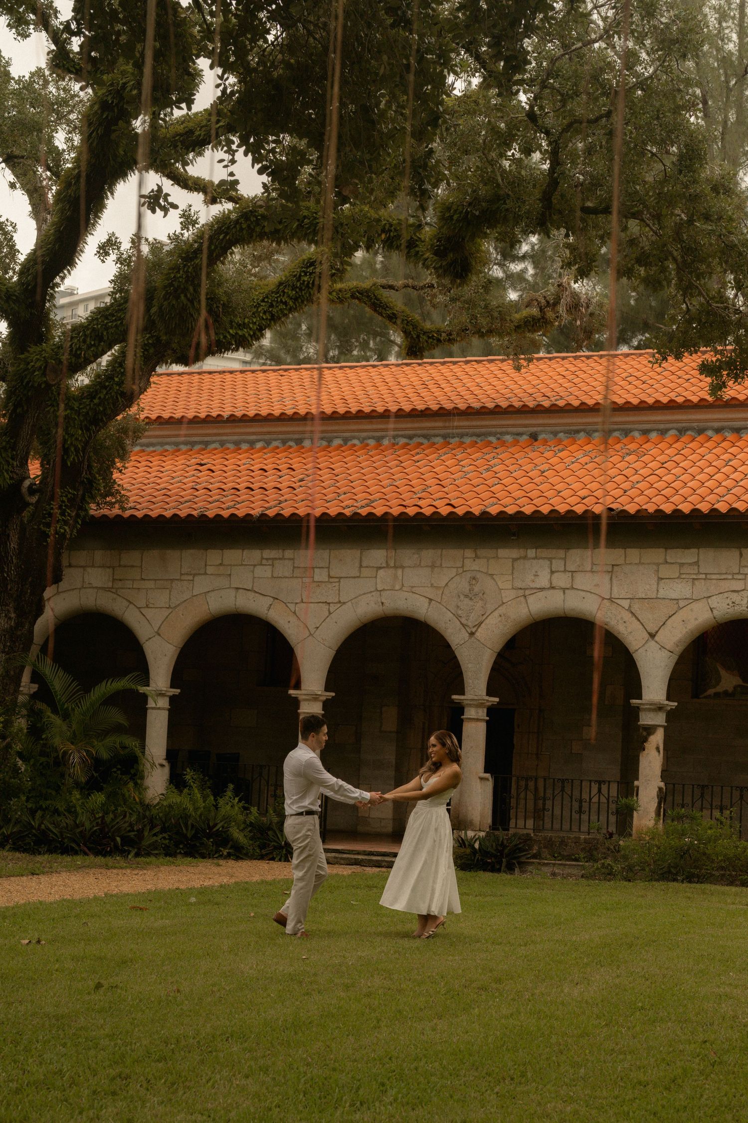 Couple holds hands and dances on a green lawn before a Spanish-style stone arcade with a terracotta roof and an oak tree.