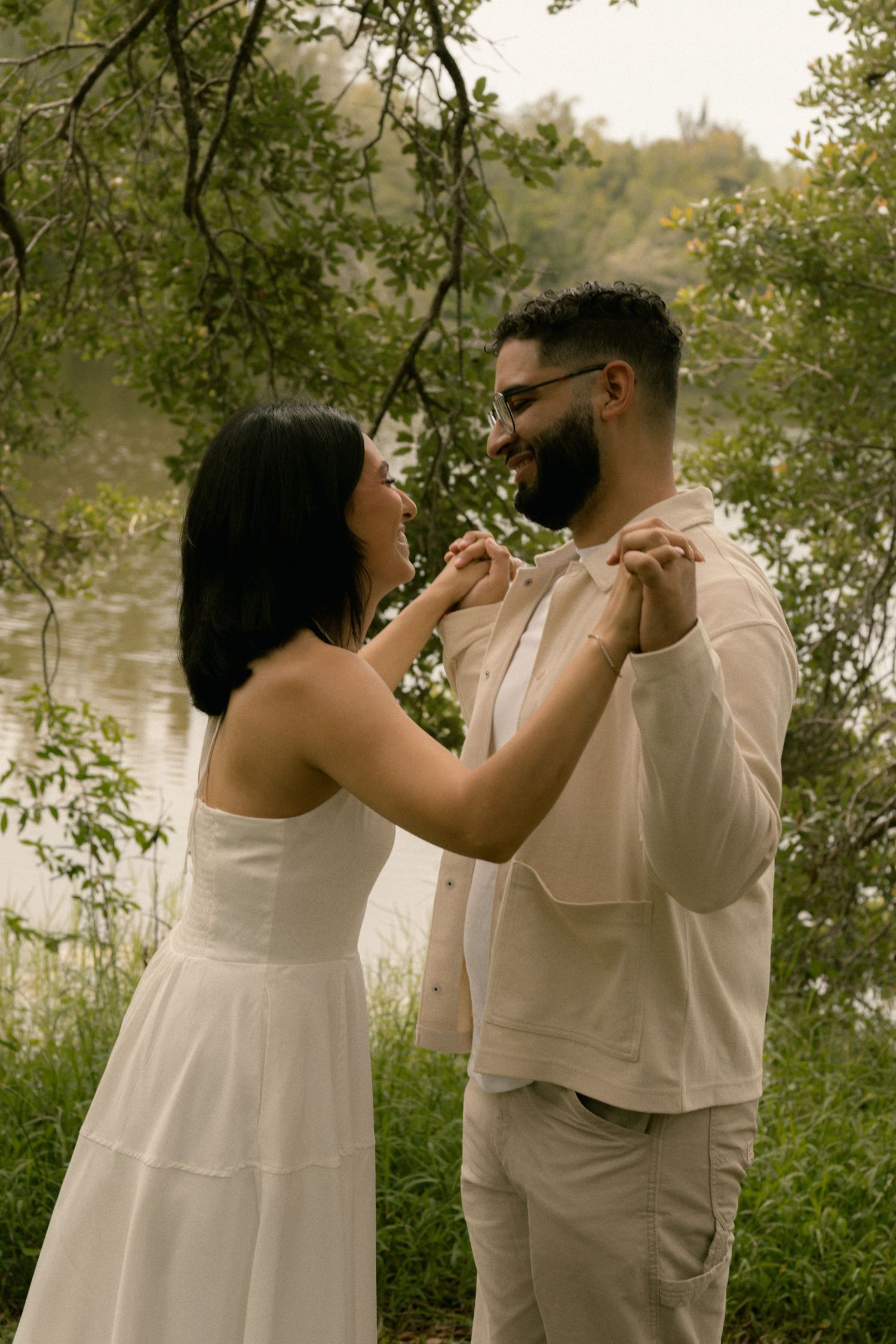 Smiling couple holds hands near a serene lakeside surrounded by lush green foliage.