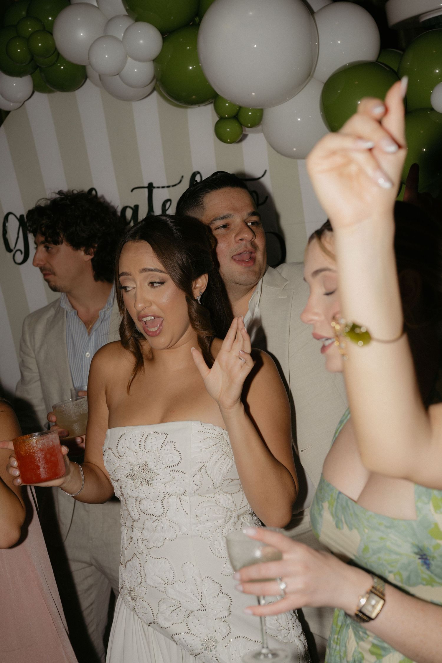 A bride in a white floral gown dances joyfully at a wedding reception with green and white balloon decorations.