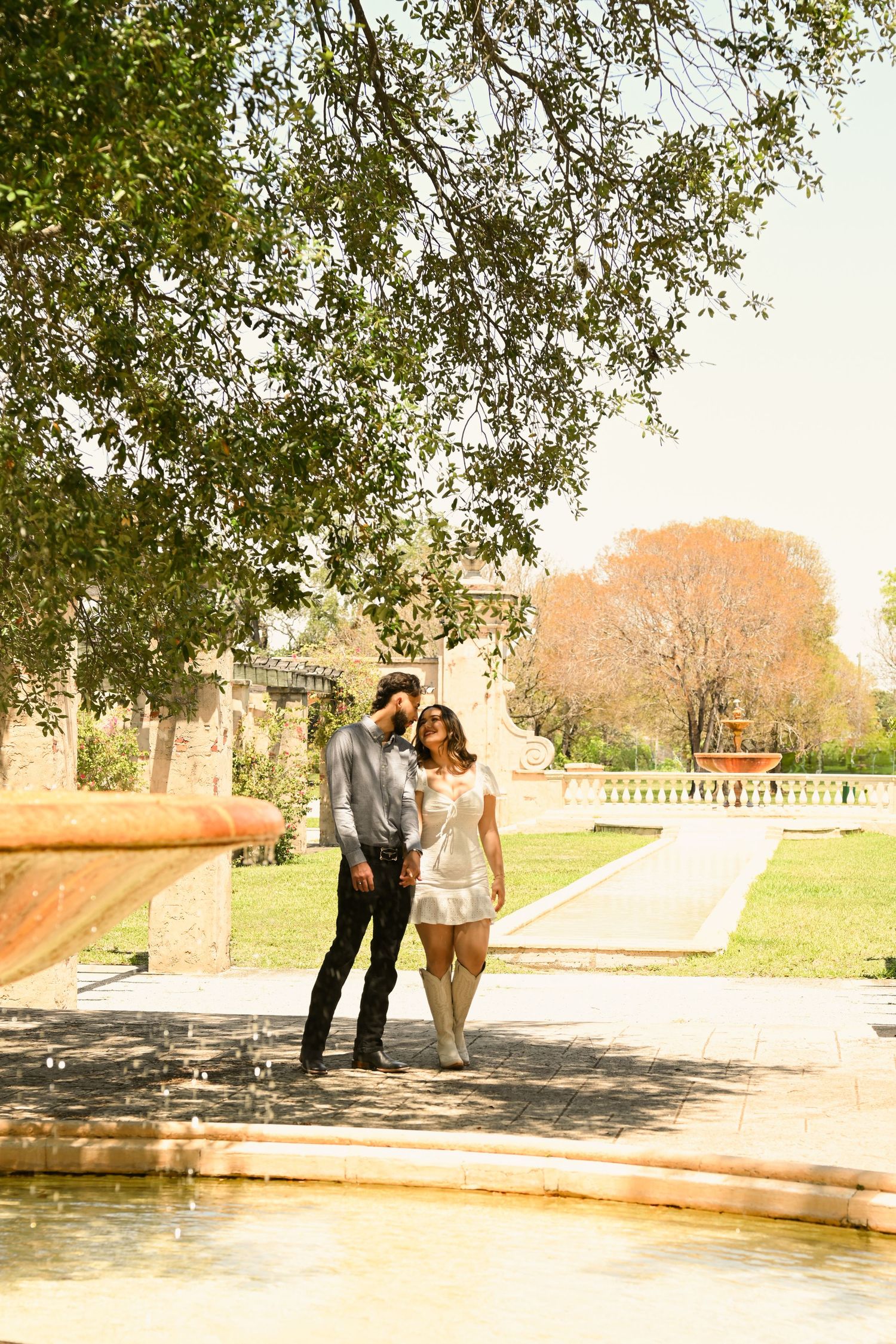 A couple poses together near a decorative fountain in a manicured garden setting.