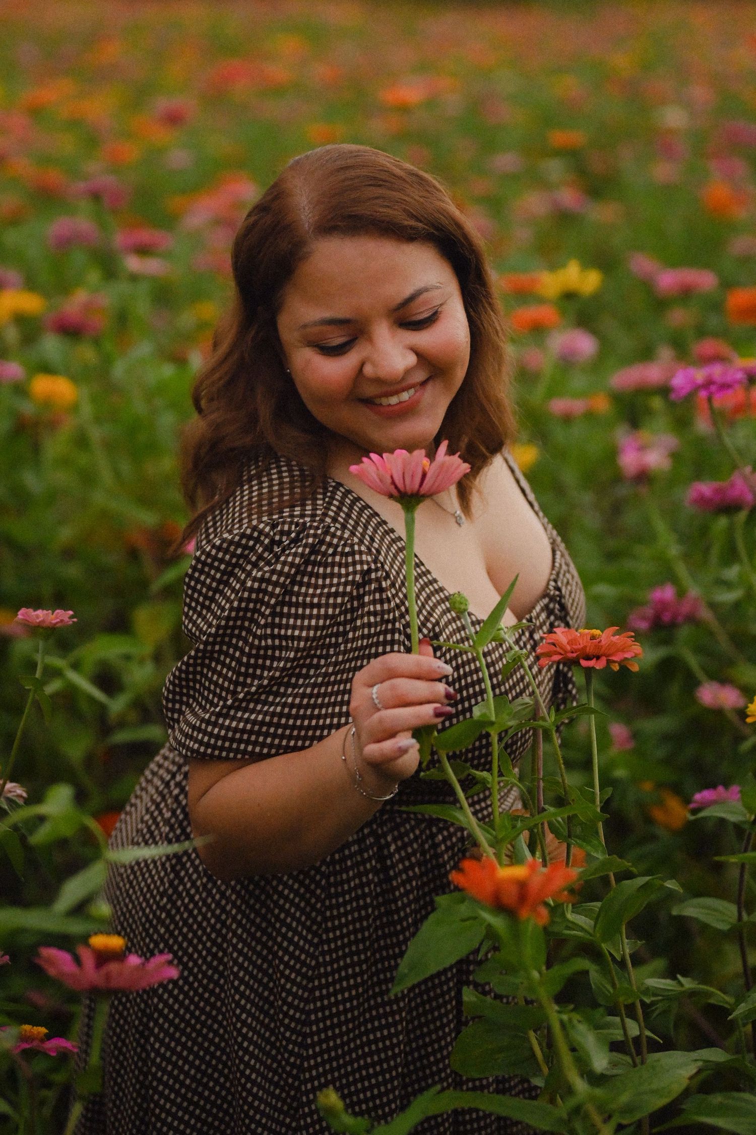 Someone in a polka dot dress smiles joyfully while holding a pink zinnia flower in a colorful summer garden.