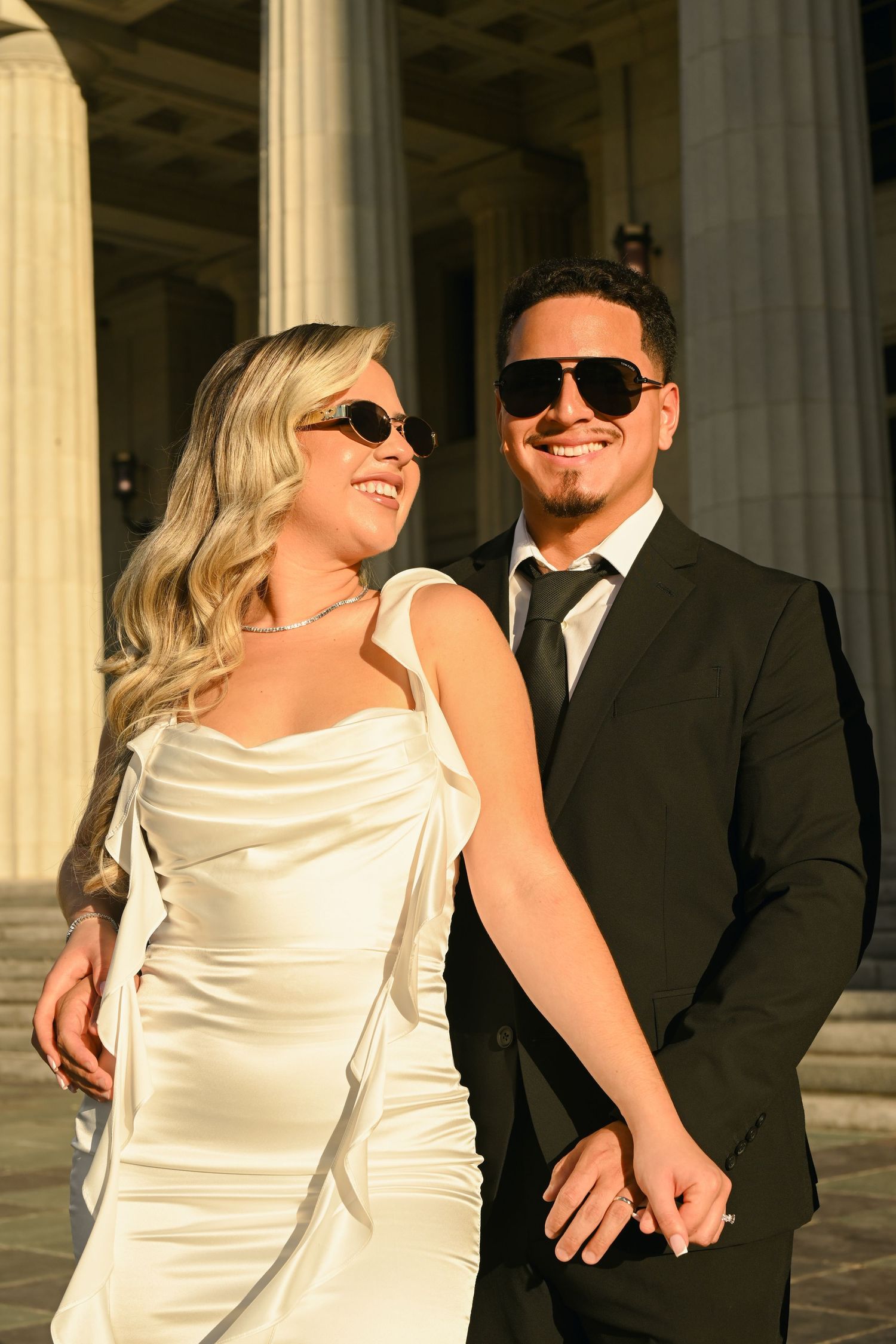 Newlywed couple in sunglasses share a romantic moment outside a courthouse building.