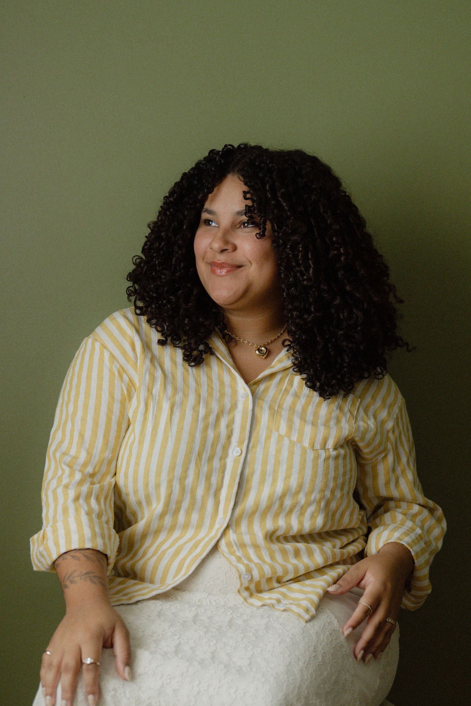 Woman with curly hair smiling in yellow striped shirt seated against olive green wall.
