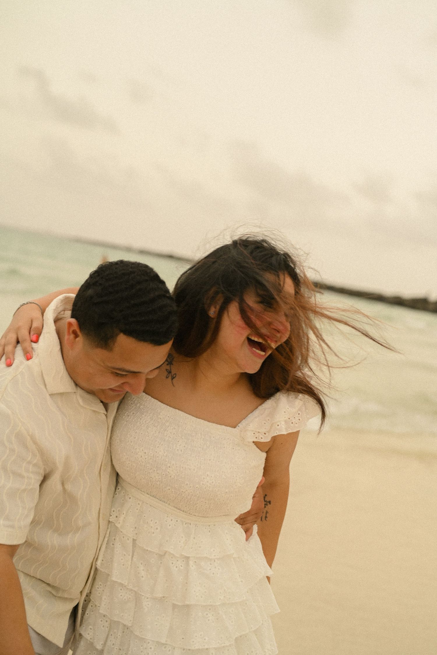 A couple in white attire shares an intimate moment while dancing on the beach at dusk.