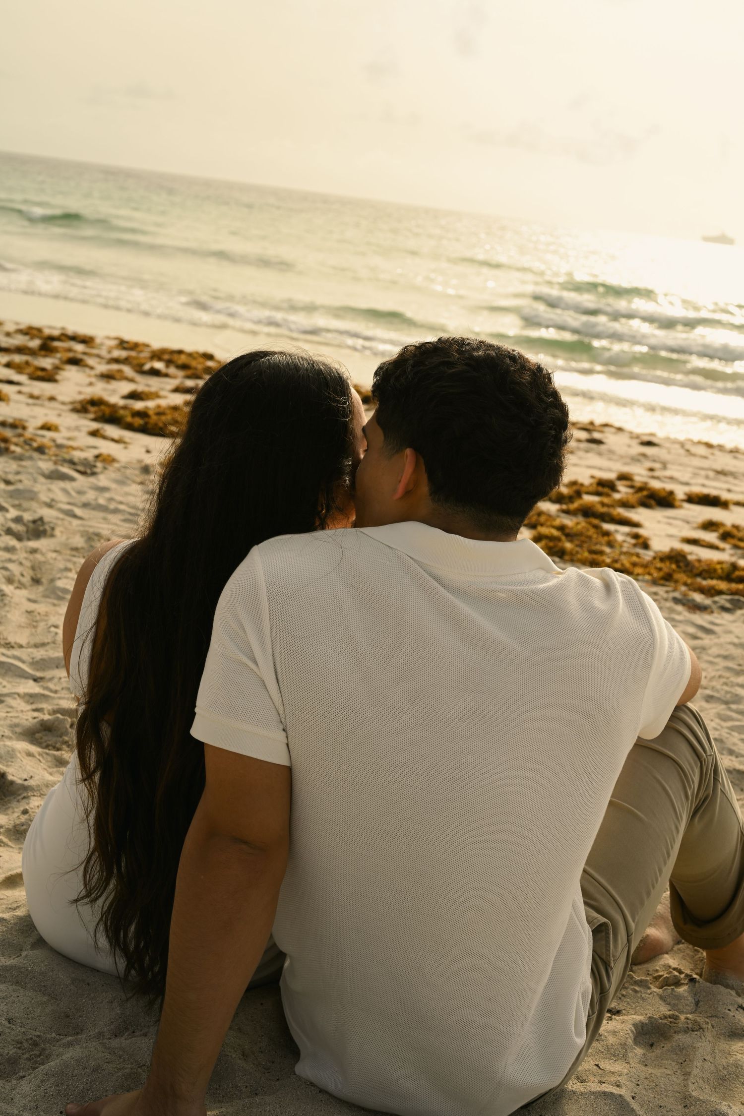A couple sits close together on the beach watching the ocean at sunset.