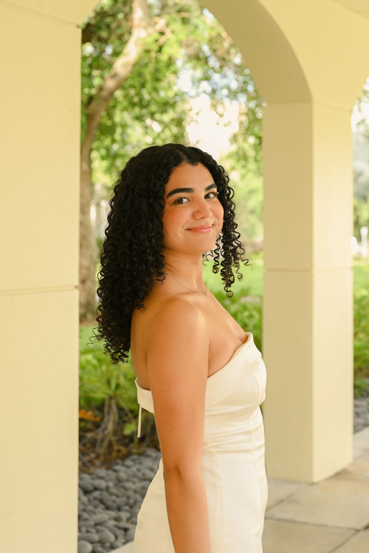 A person in a white dress poses in an elegant colonnade with natural lighting.