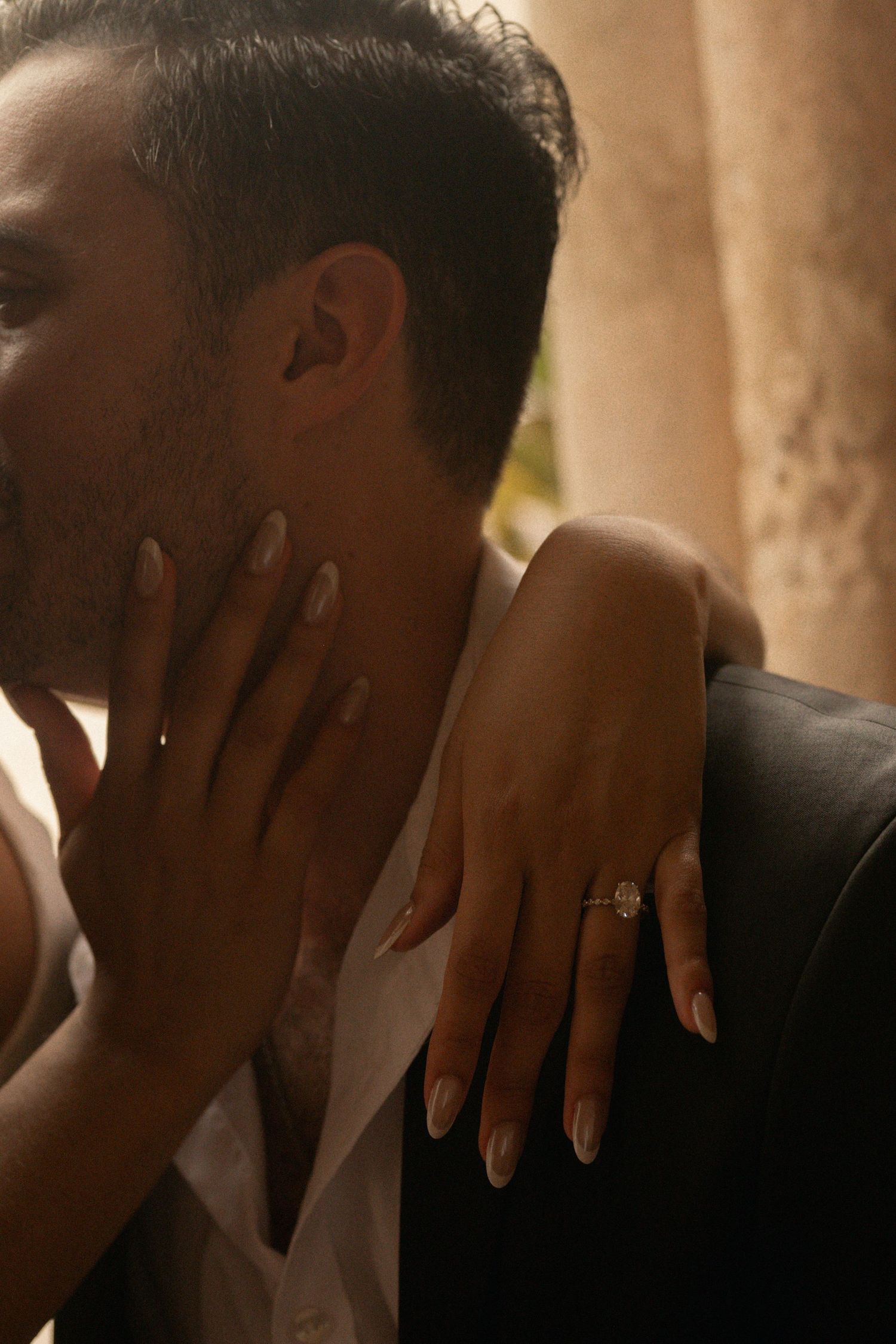 Woman's hand with a diamond engagement ring resting on a man's shoulder as her other hand touches his face in an intimate close-up.