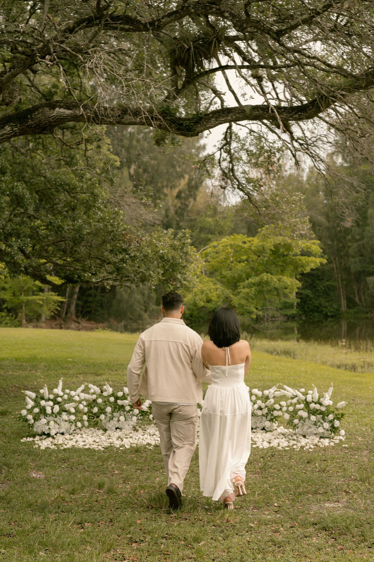 Couple walks hand-in-hand toward a floral arrangement in a lush green park during an outdoor proposal.