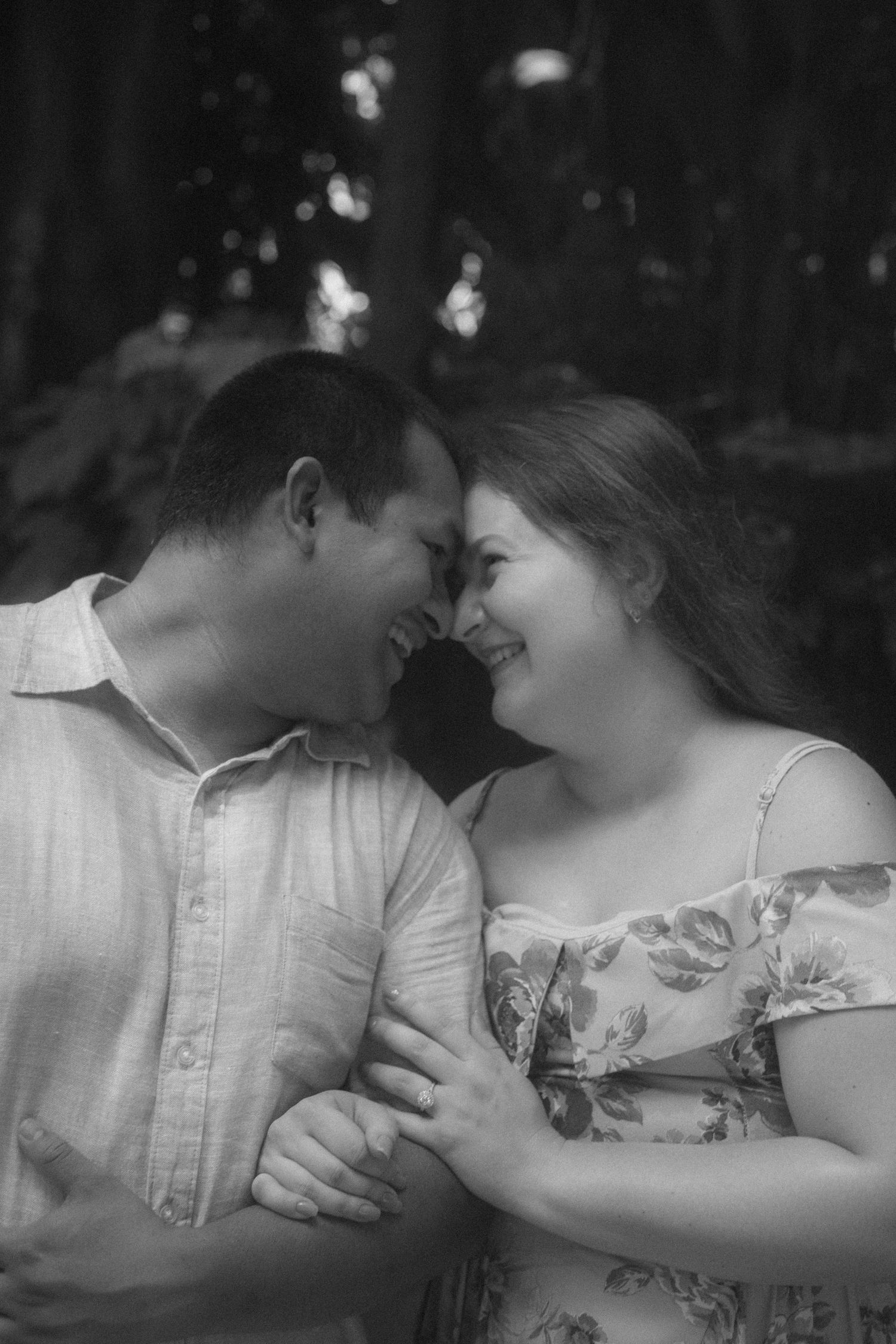 Black and white portrait of a smiling couple touching foreheads tenderly in a forest setting.