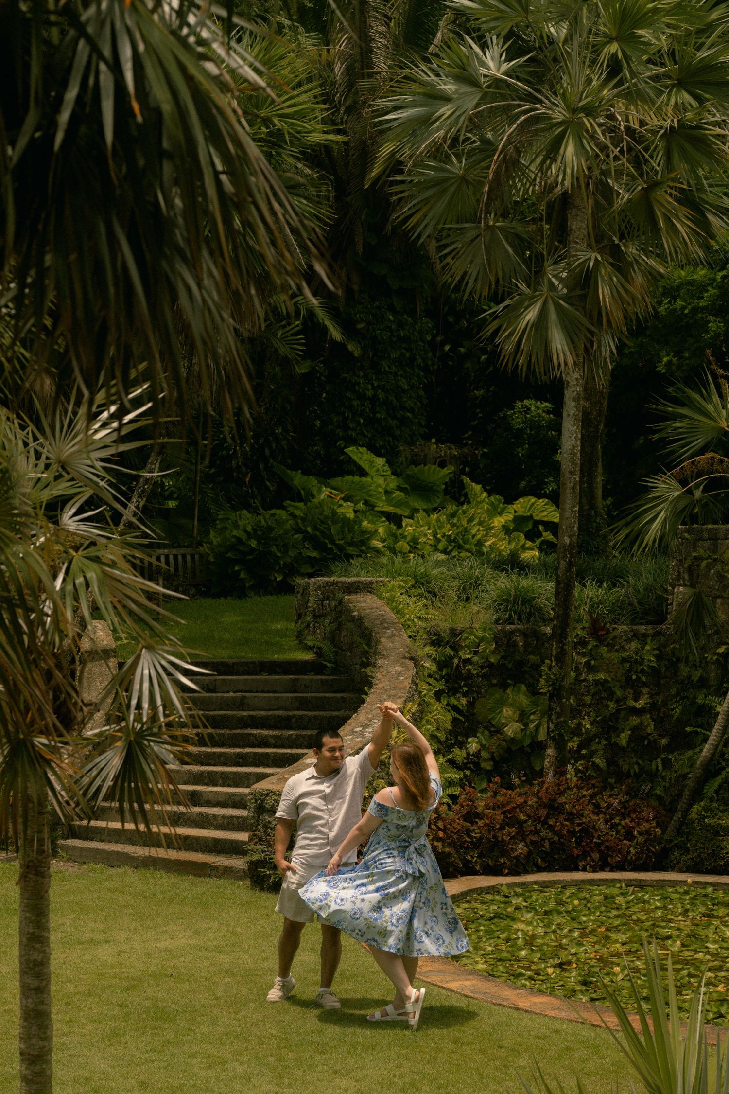 Couple dancing joyfully on a lush green lawn surrounded by tropical palm trees and stone steps in a garden.