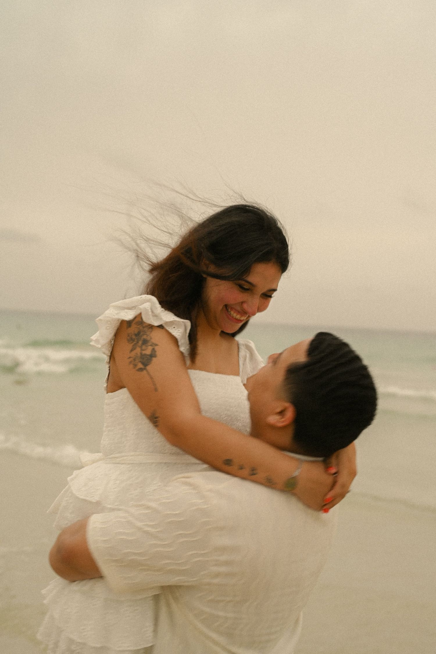 A couple in white clothing share romantic embraces on a cloudy beach with ocean waves in the background.