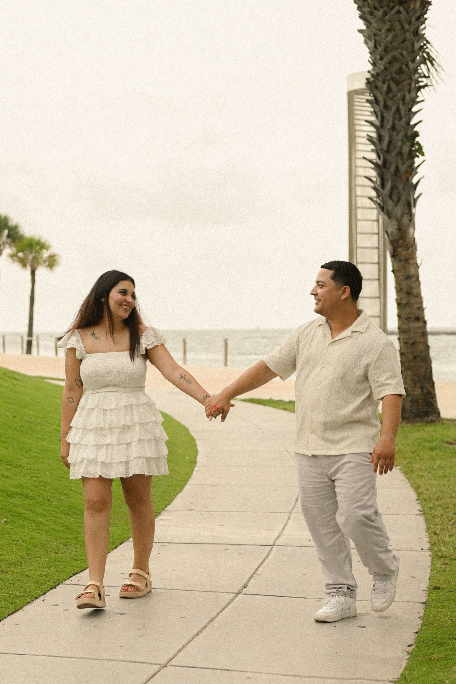 A couple in white outfits holds hands while walking along a palm-lined pathway.