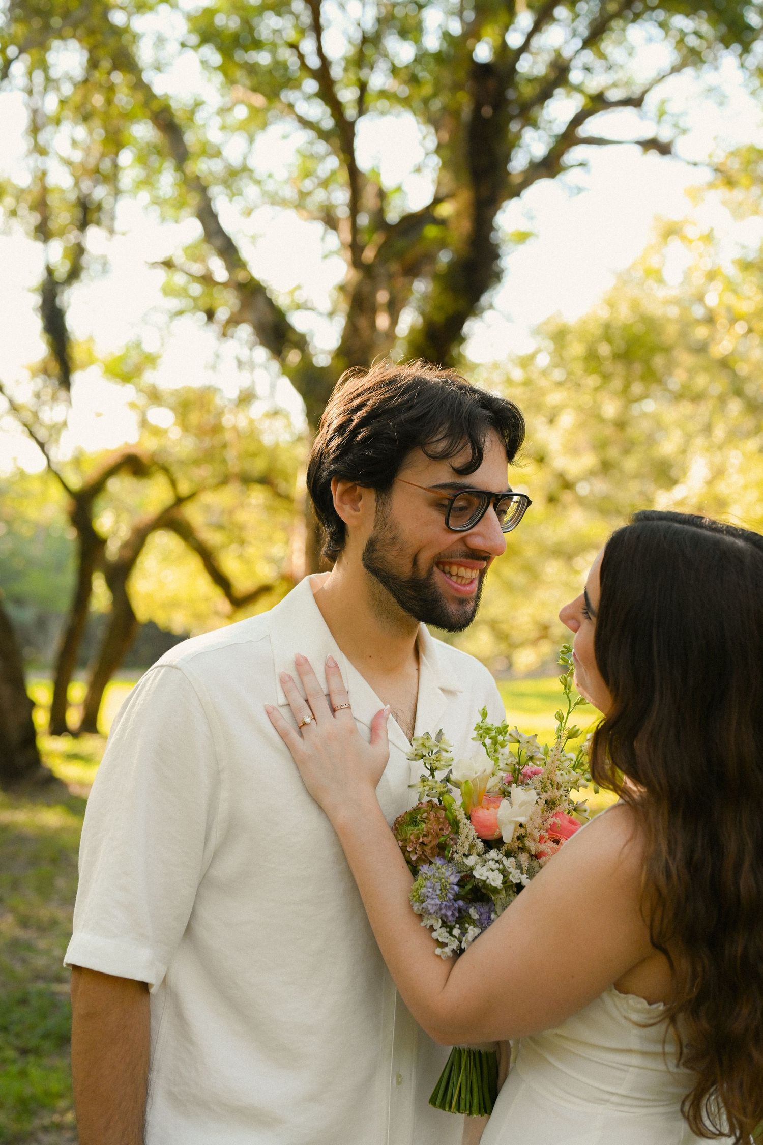 A couple shares a romantic moment in a sunlit outdoor setting with trees and flowers in the background.