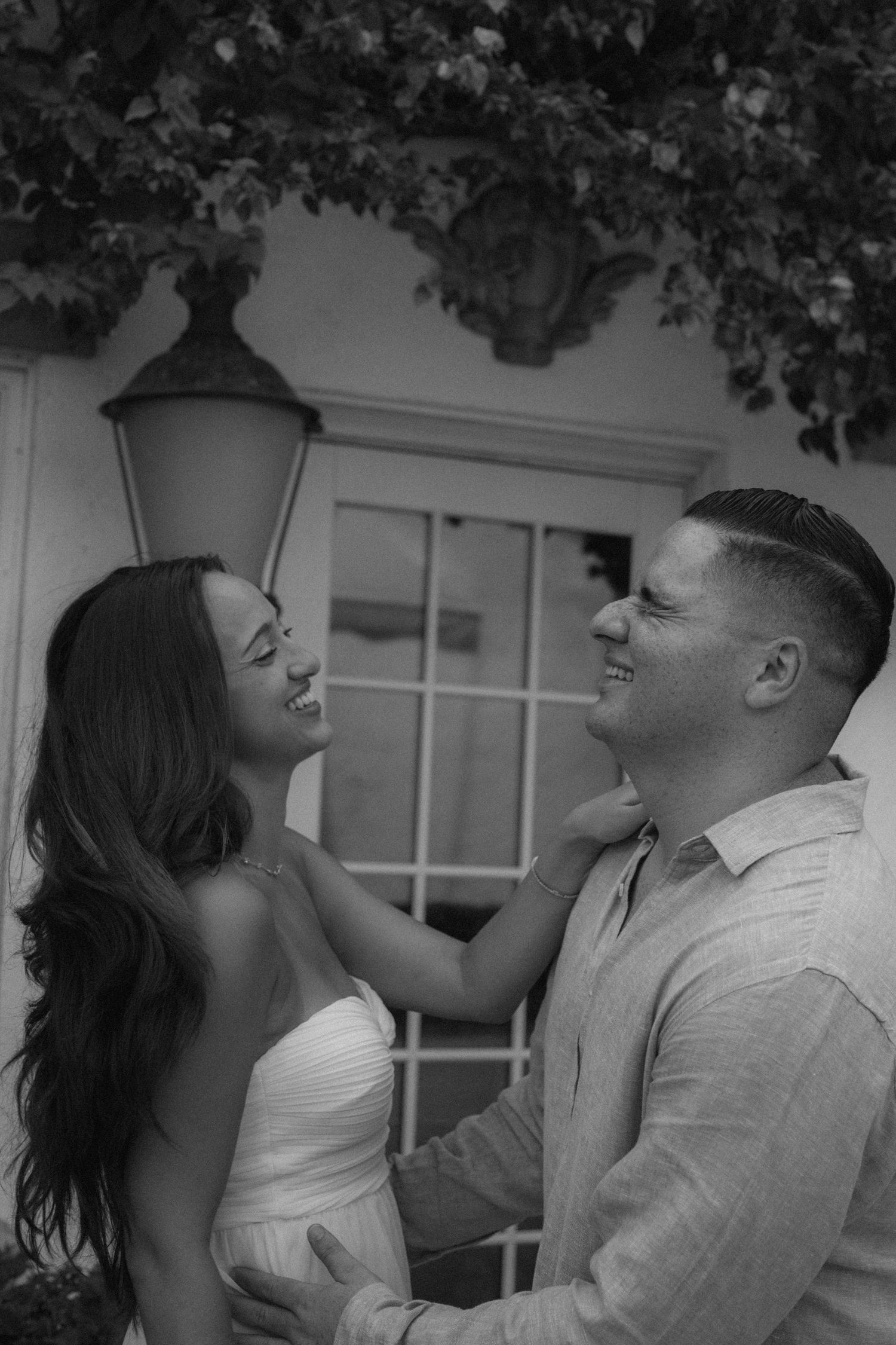 Black and white photo of a smiling couple in formal wear, woman in white dress touching mans collar outdoors.