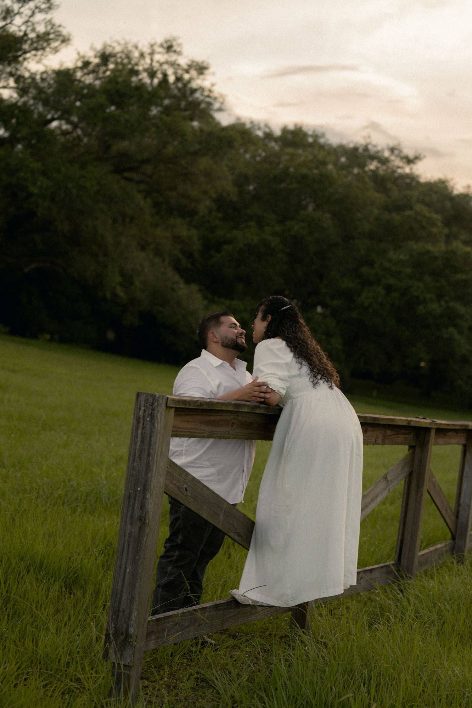 Couple in white embracing by rustic wooden fence in lush green field at sunset.