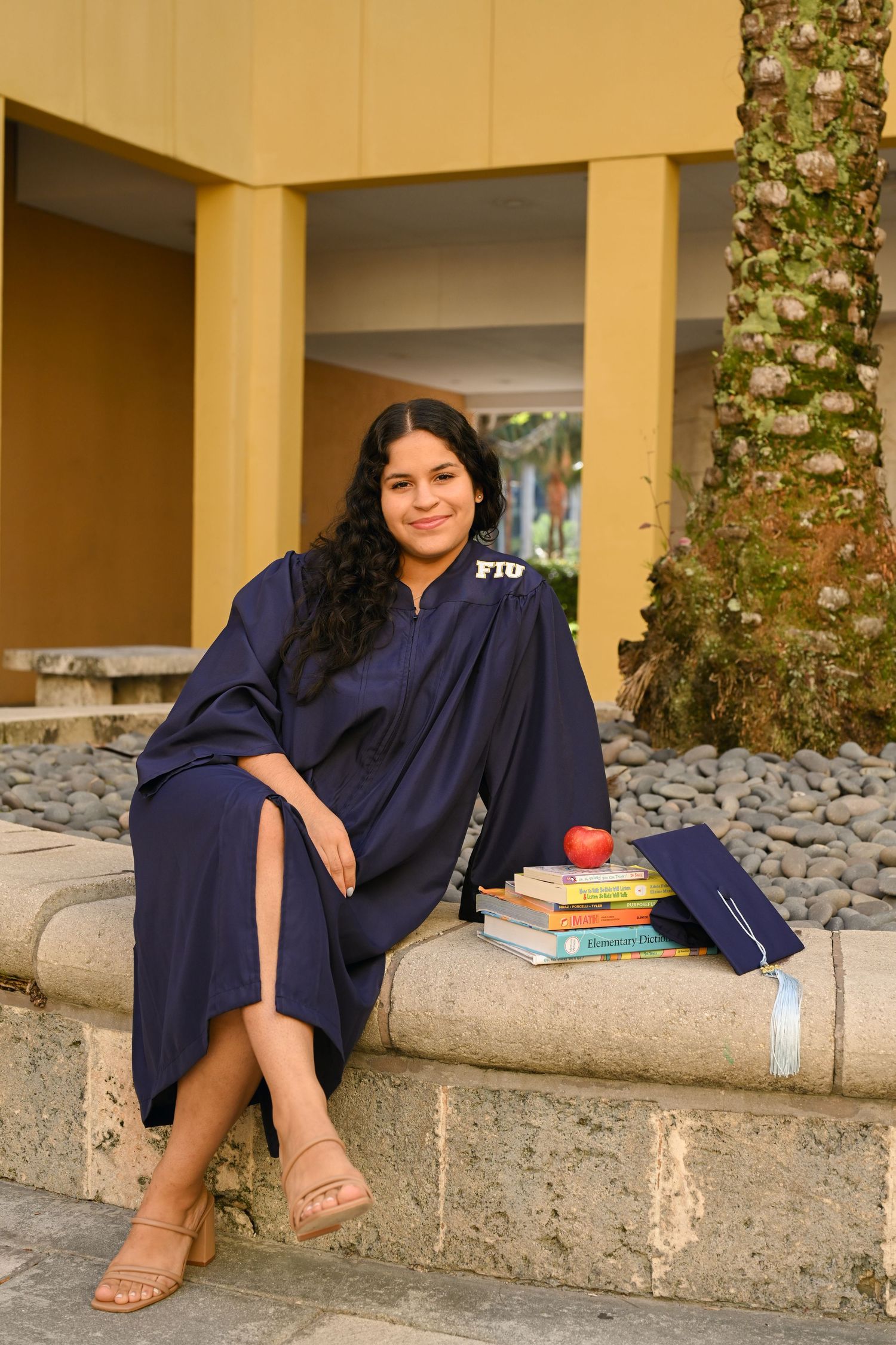 Graduate in navy regalia poses with books and apple on concrete ledge.