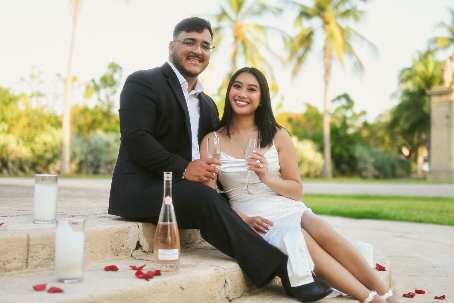 Couple sharing champagne and intimate moments on stone steps with palm trees in background.