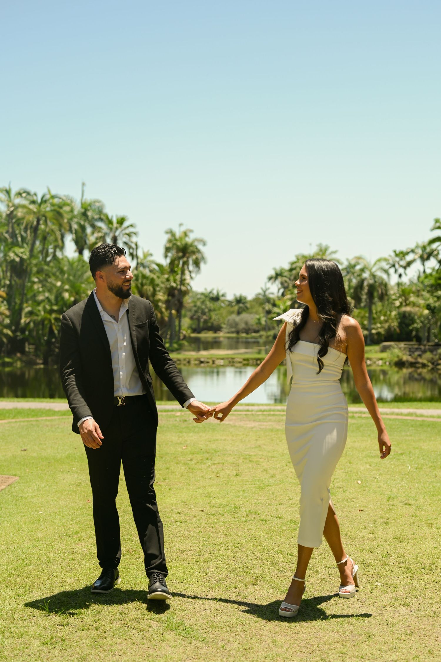 A couple playfully dances on a sunny lawn with palm trees and a lake in the background.