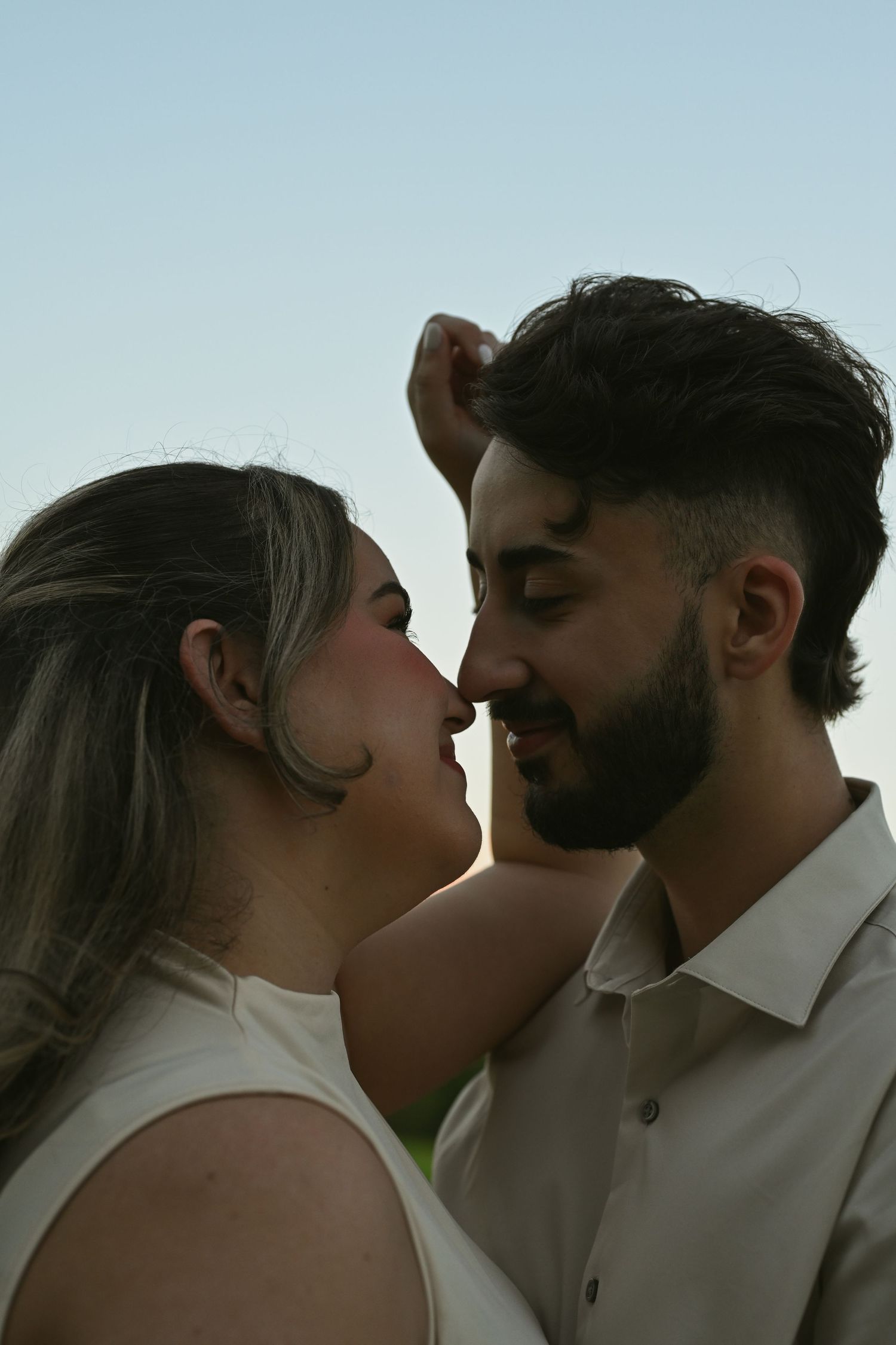 Romantic couple sharing an intimate moment, nearly kissing outdoors against a soft blue sky backdrop.