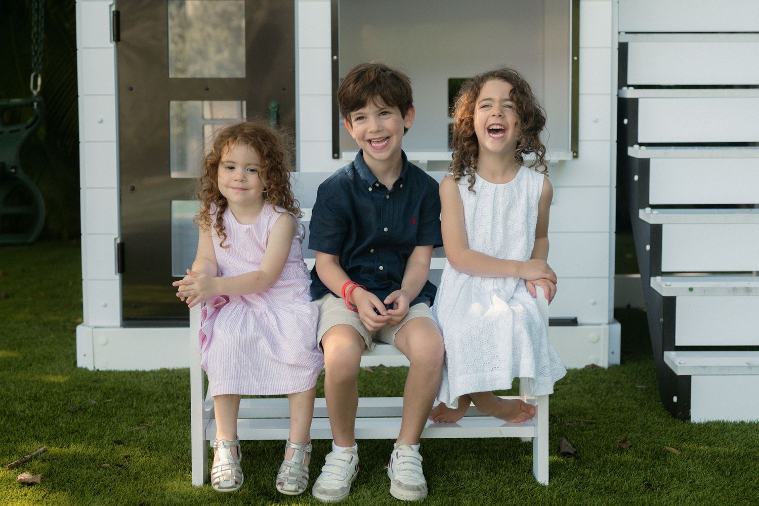 Three smiling young children sit together on a small white bench in front of a white playhouse on a sunny green lawn.