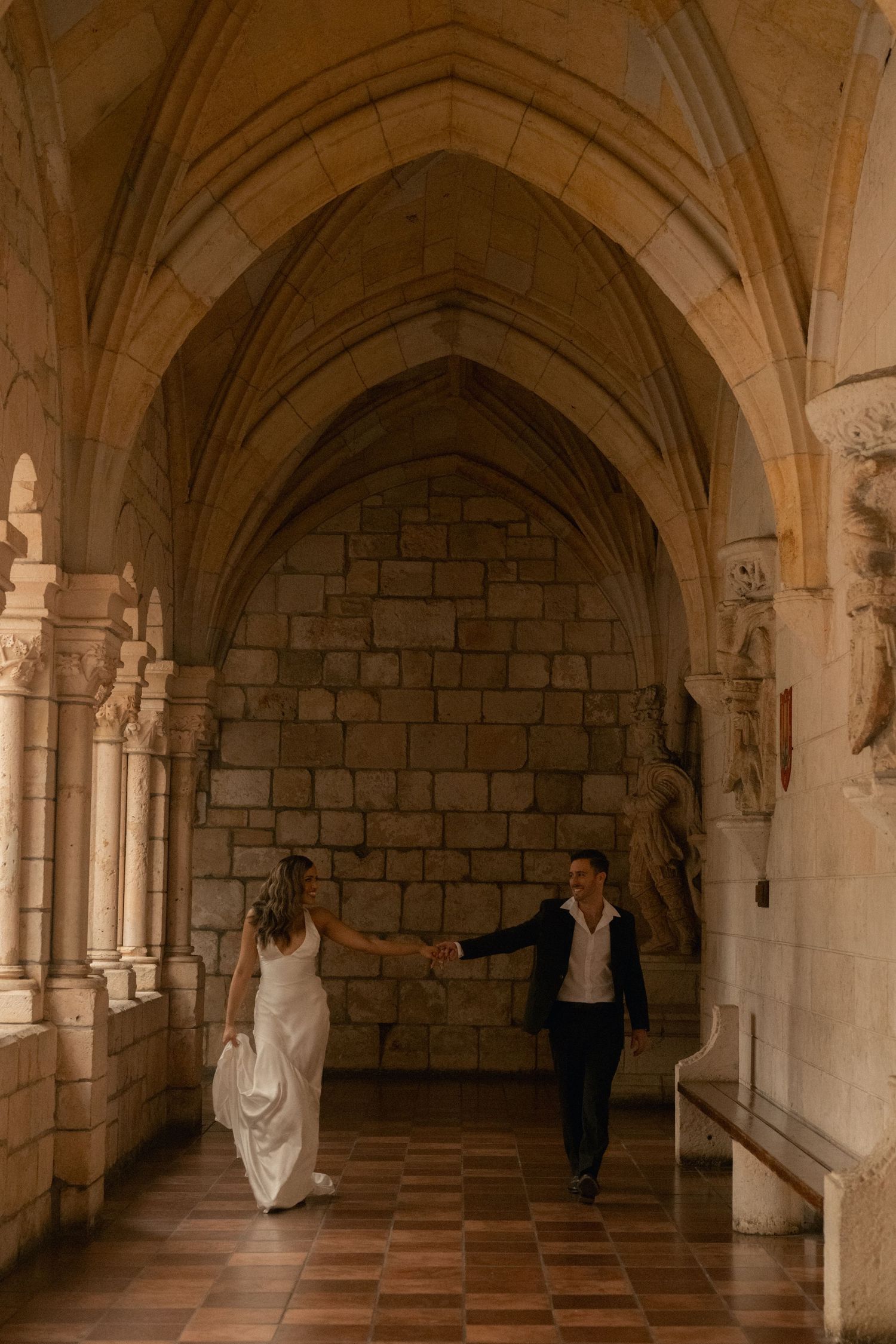 Bride in a white gown holds hands with a groom in a dark blazer while walking through a historic stone cloister with vaulted arches.
