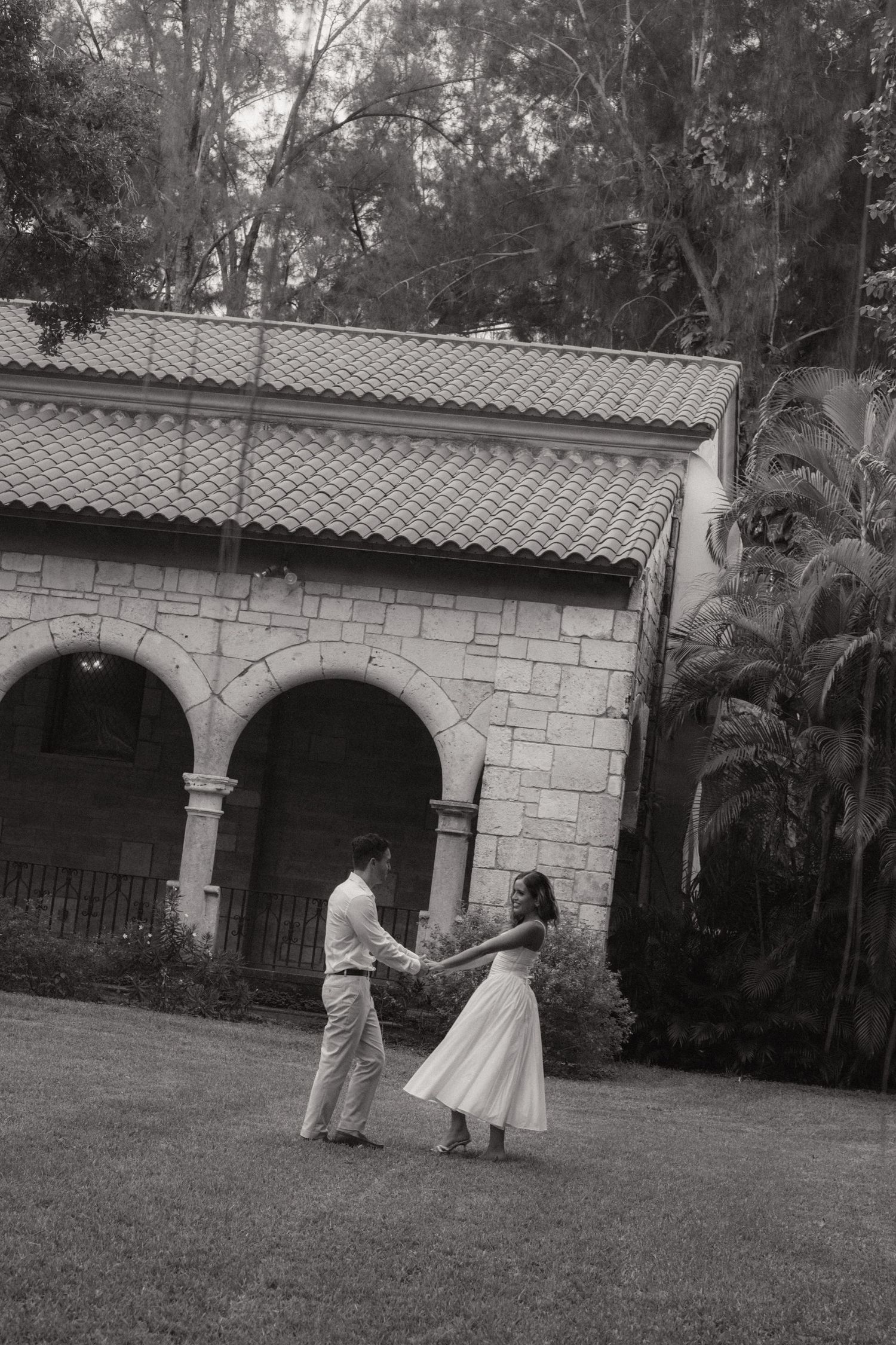 Black and white photo of a couple holding hands and dancing on a lawn before a stone arcade with arches and a tiled roof.