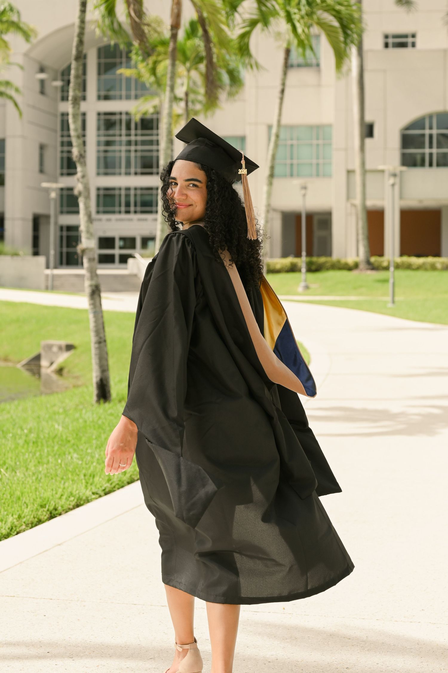 Graduate in black cap and gown poses joyfully on a sunny campus walkway with palm trees.