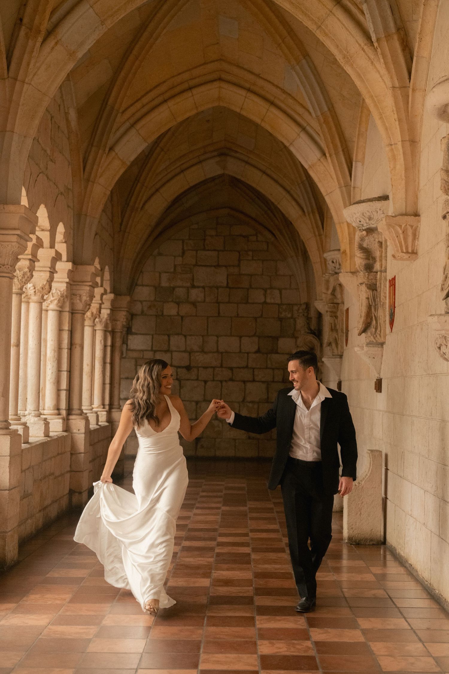 Bride in a flowing white gown and groom in a dark suit hold hands and walk through a historic stone cloister with arched ceilings.