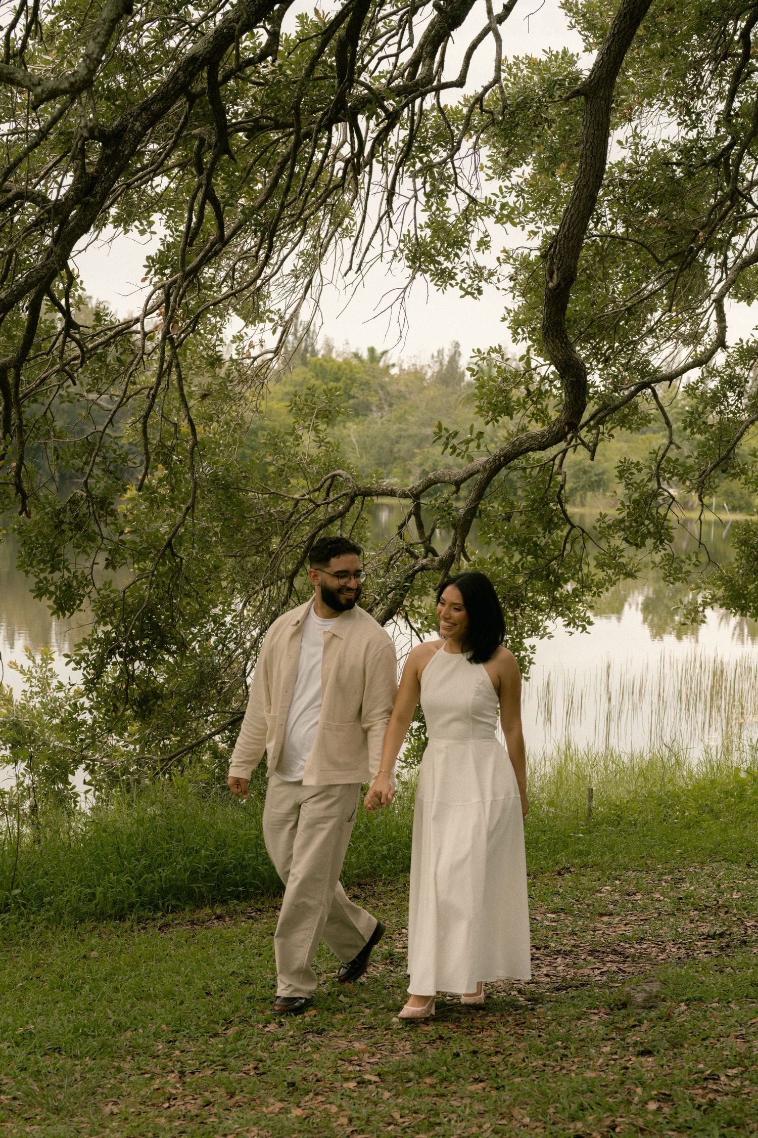 A smiling couple walks hand-in-hand near a tranquil lake surrounded by lush green trees and hanging branches.