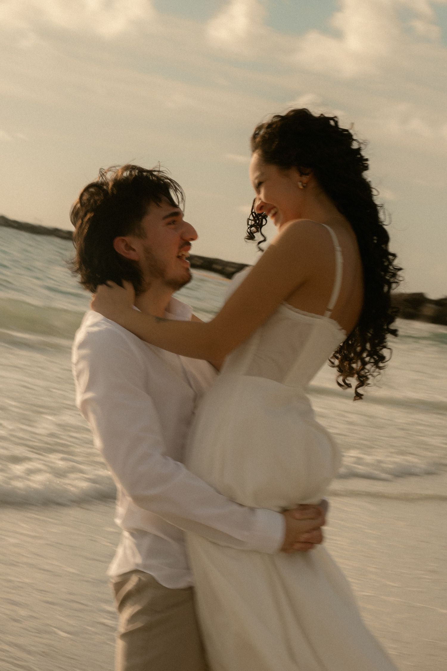 A joyful couple embraces on a beach at sunset, the woman in a white dress and man in white shirt.