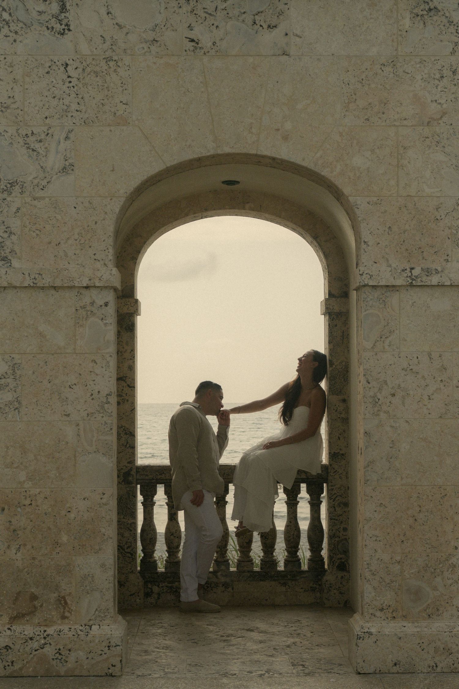 Couple in wedding attire sharing a romantic moment framed by a stone arch overlooking the ocean at sunset.