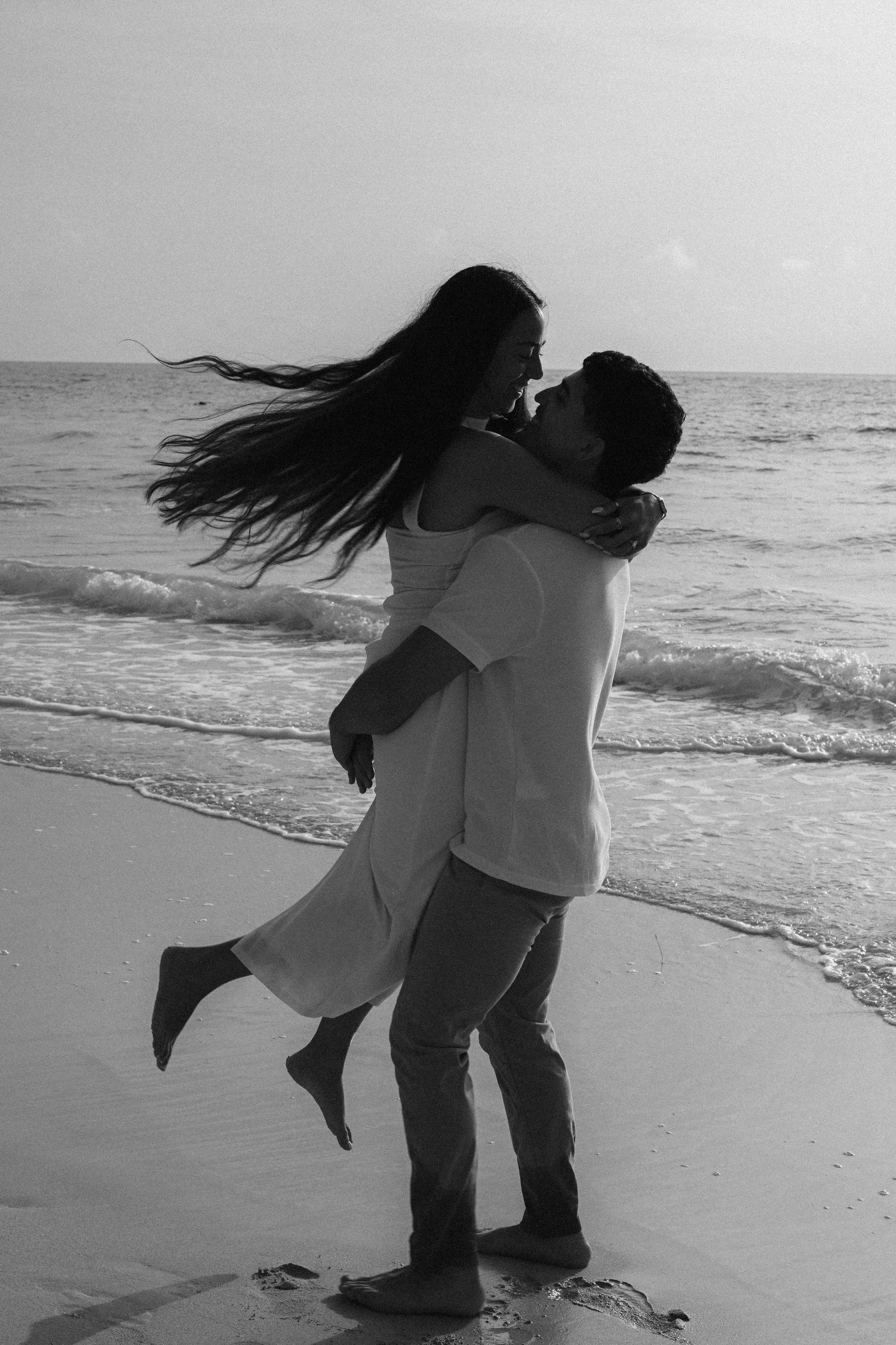 Black and white photo of a couple embracing on a beach, man lifting woman as her hair blows in the wind.