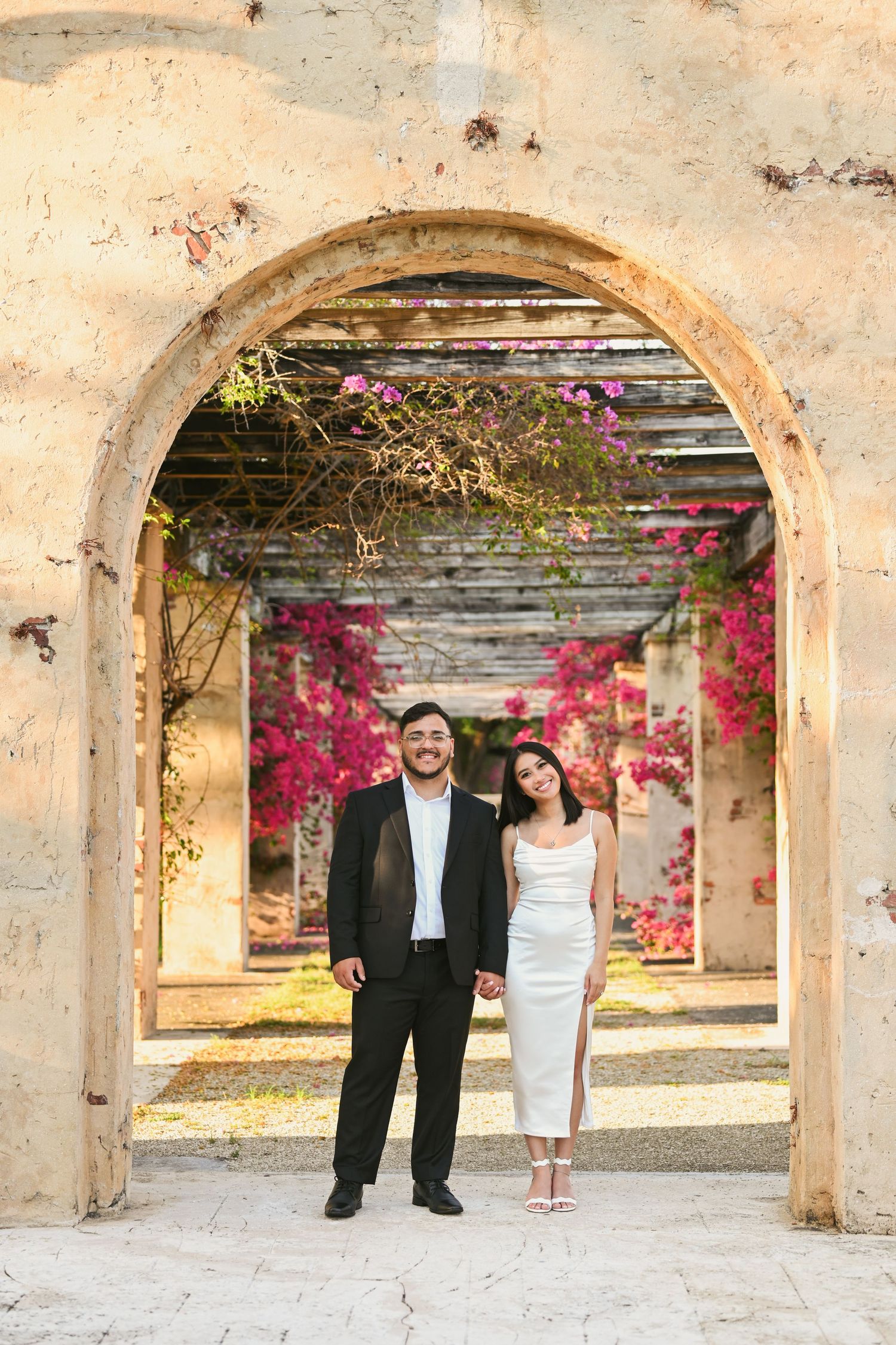 Couple standing under stone archways decorated with pink bougainvillea flowers.