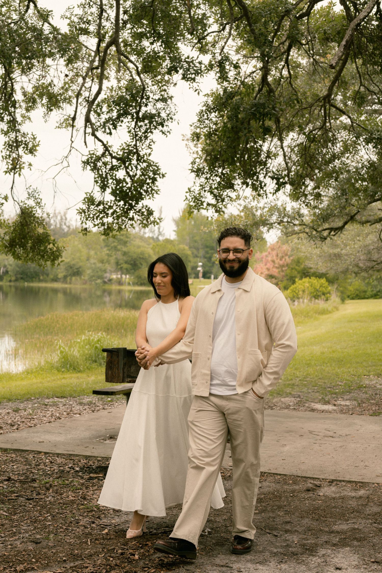 Couple walking together in a lush park near a pond, woman in white dress and man in beige outfit.