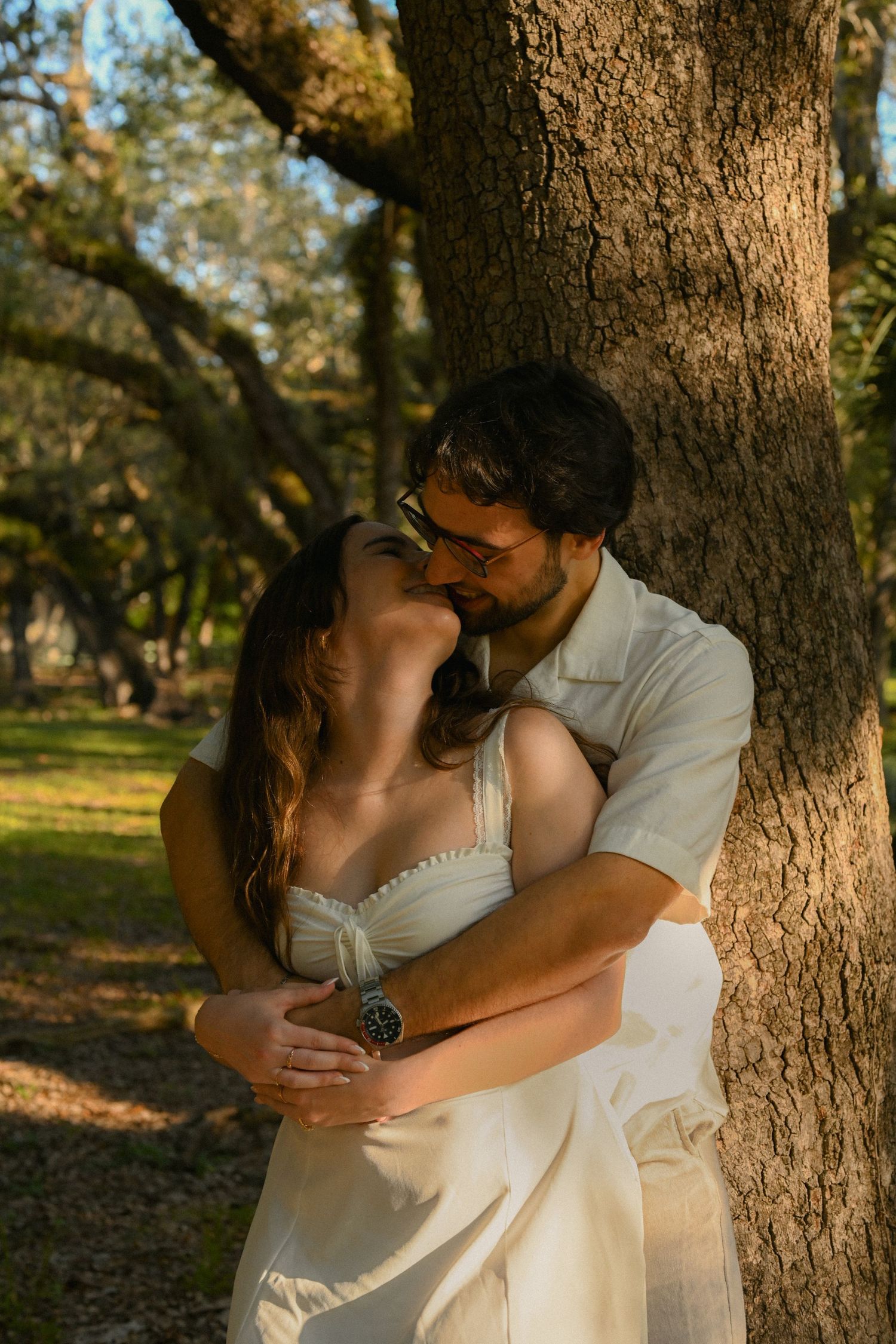 A romantic couple embraces tenderly against a tree trunk in a sunlit park setting during golden hour.