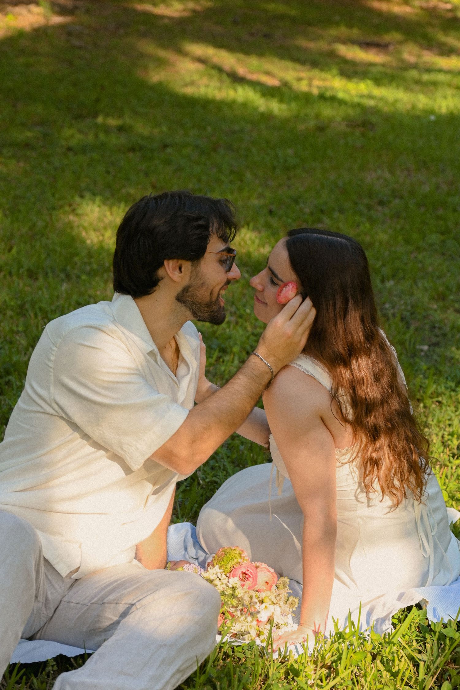 A couple in white outfits share romantic moments while sitting on grass with flowers in a sunlit outdoor setting.