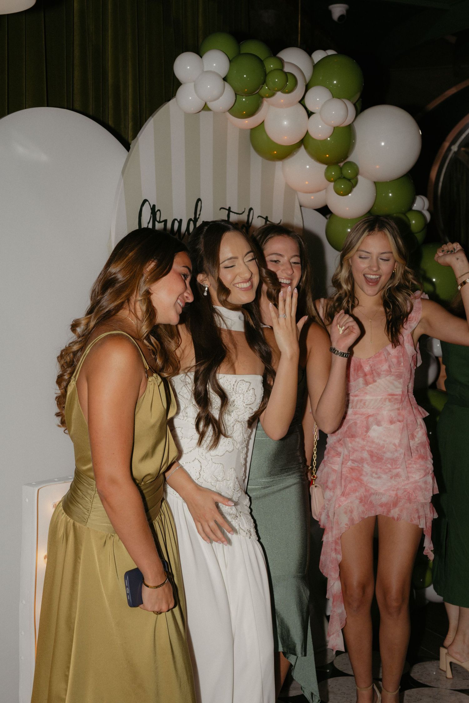Four young women in formal dresses celebrate at a graduation party with green and white balloon decorations.