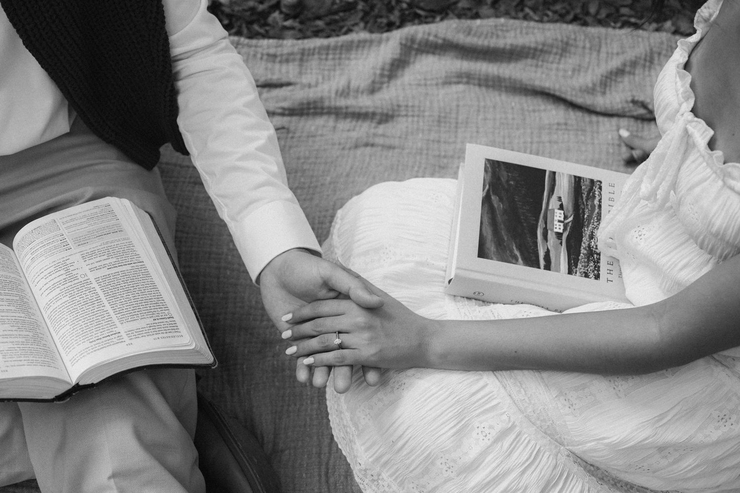 Bride and groom holding hands with open Bible and wedding photo on blanket in black and white.