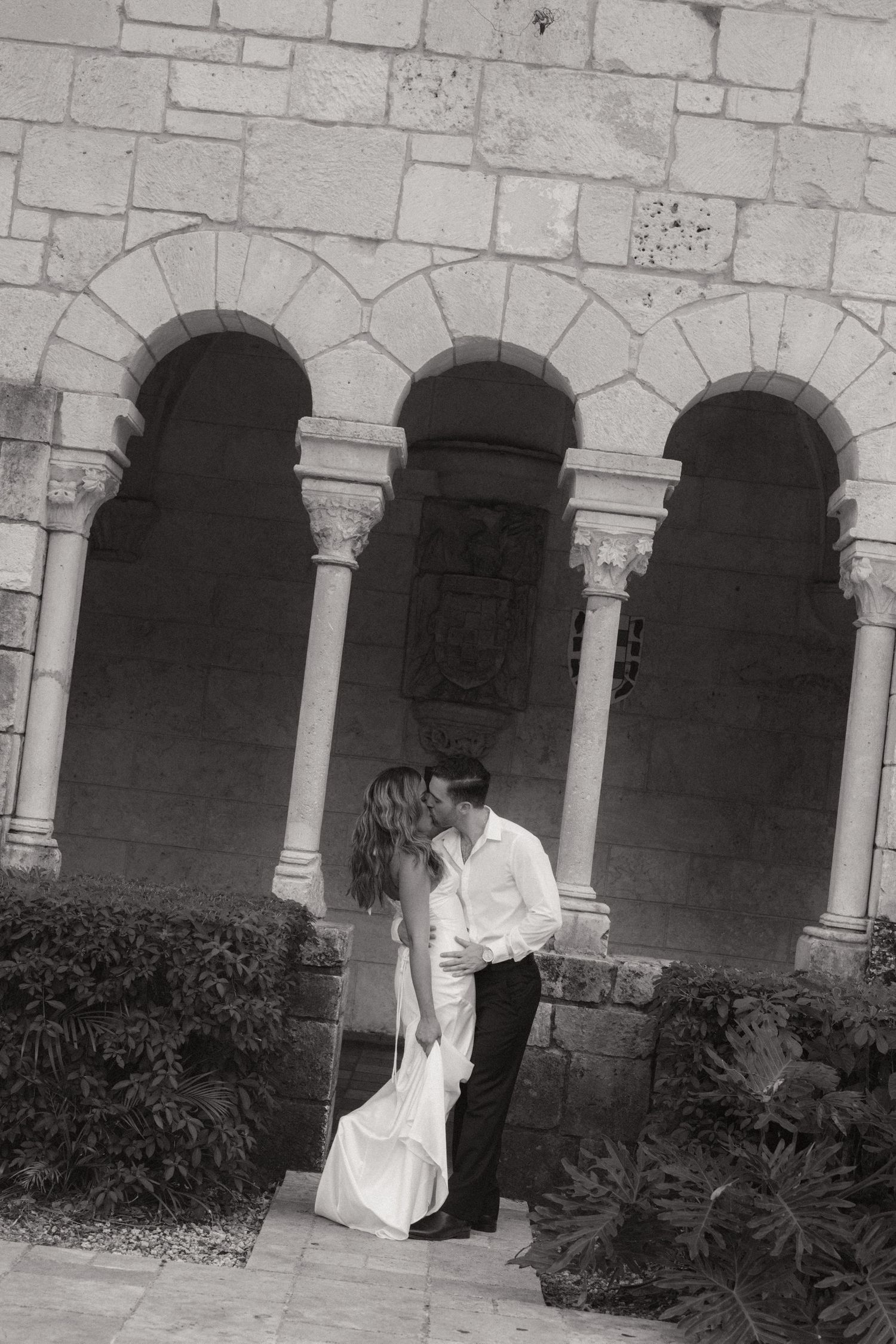 Black and white photo of a bride in a flowing gown kissing her groom beneath historic stone arches with carved columns.