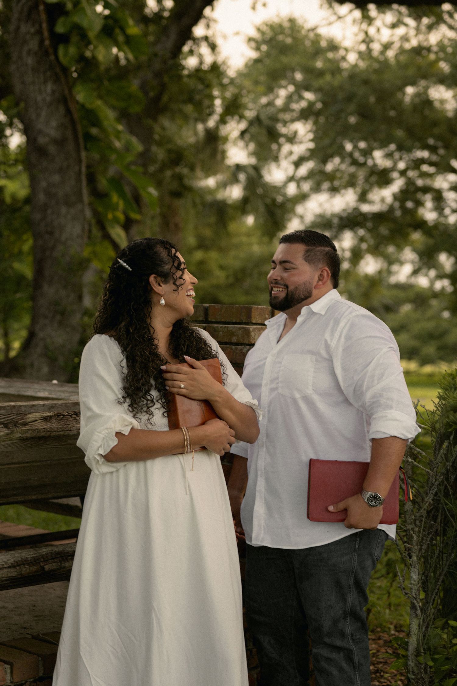 Smiling couple sharing a tender moment outdoors, woman in white dress, man holding a red book.