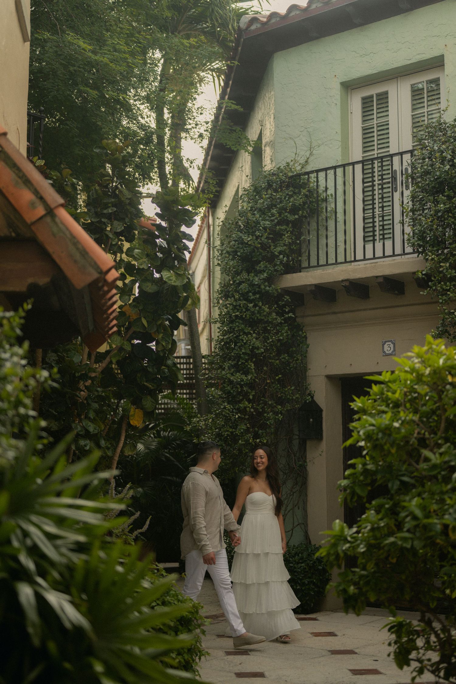 Couple in wedding attire walking hand-in-hand through a lush courtyard beside an ivy-covered building.